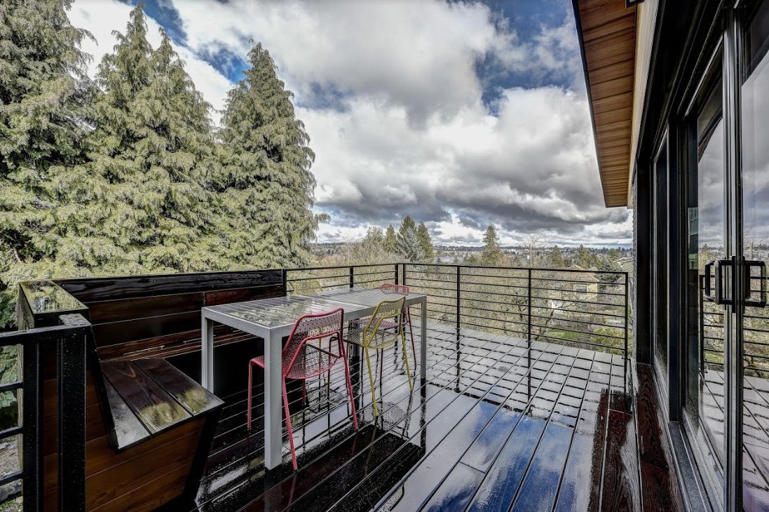 Outdoor balcony with a table and colorful chairs, overlooking a lush forest landscape under a cloudy sky.