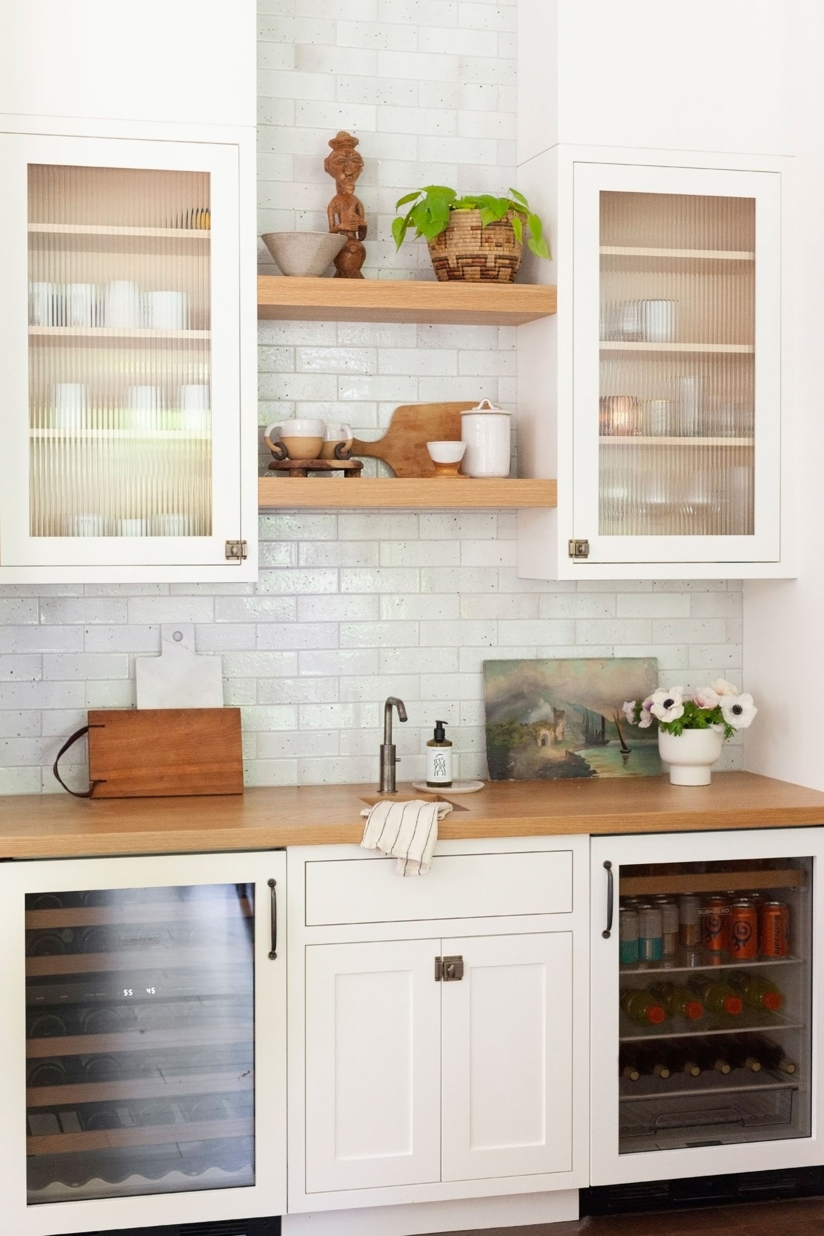 Modern kitchen with white cabinets, wooden countertops, a small sink, and decorative shelves. Upper shelves hold potted plants, a wooden sculpture, and kitchen items. Lower section features a beverage cooler and a wine fridge.