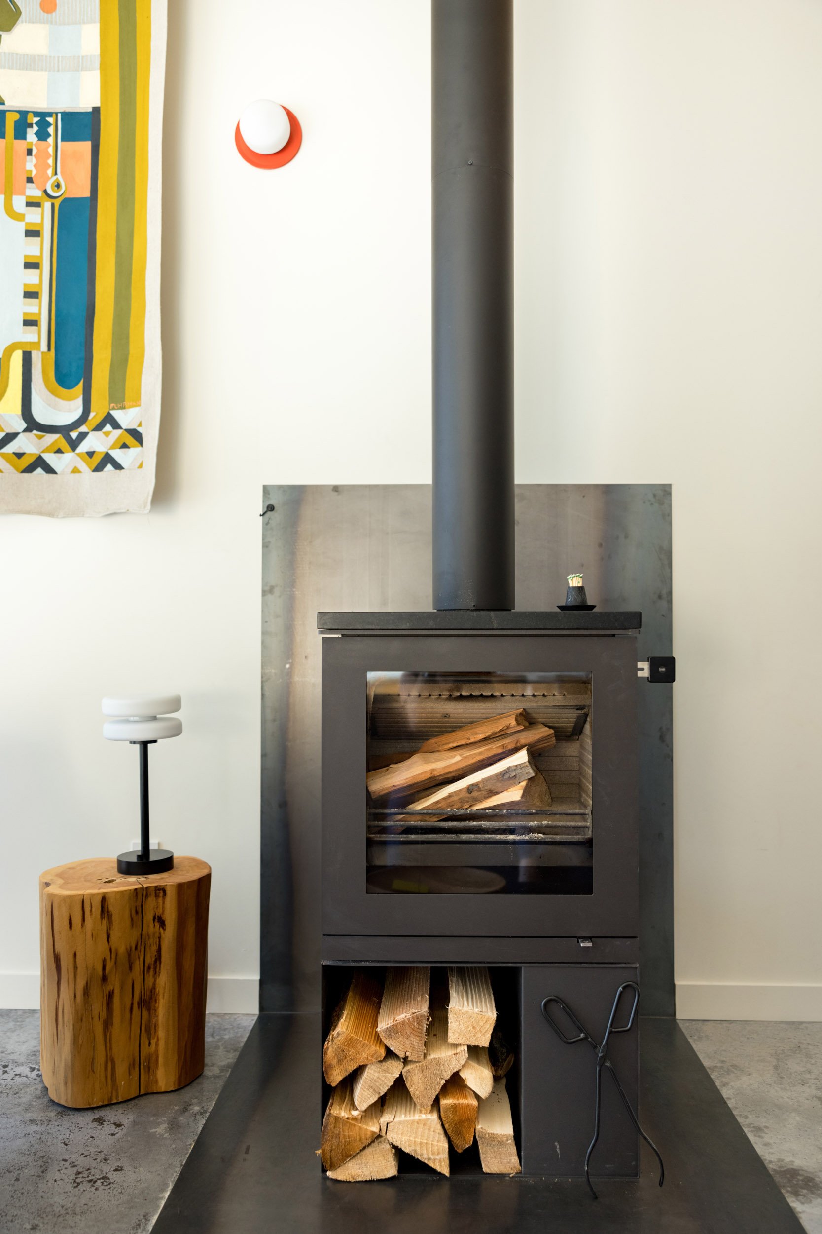 Modern wood-burning stove with chimney and stacked firewood, next to a wooden side table with a small lamp in a minimalist interior.