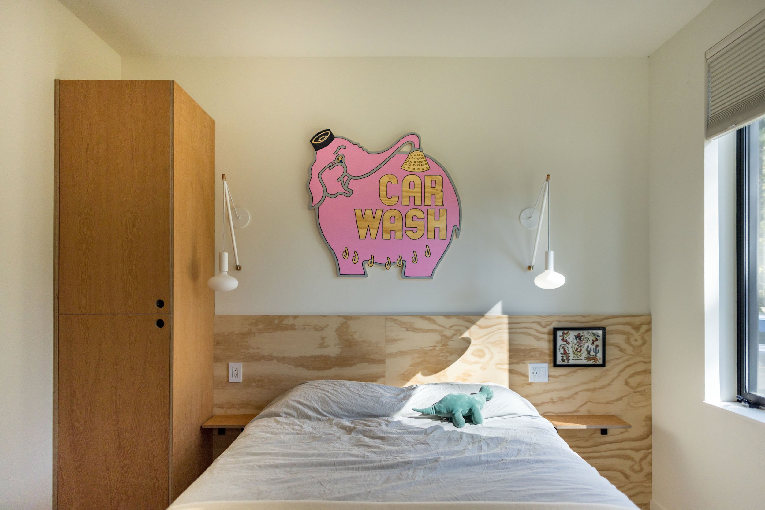 Minimalist bedroom with a bed, light wood cabinetry, wall lamps, and a playful pink elephant "Car Wash" sign above the bed.