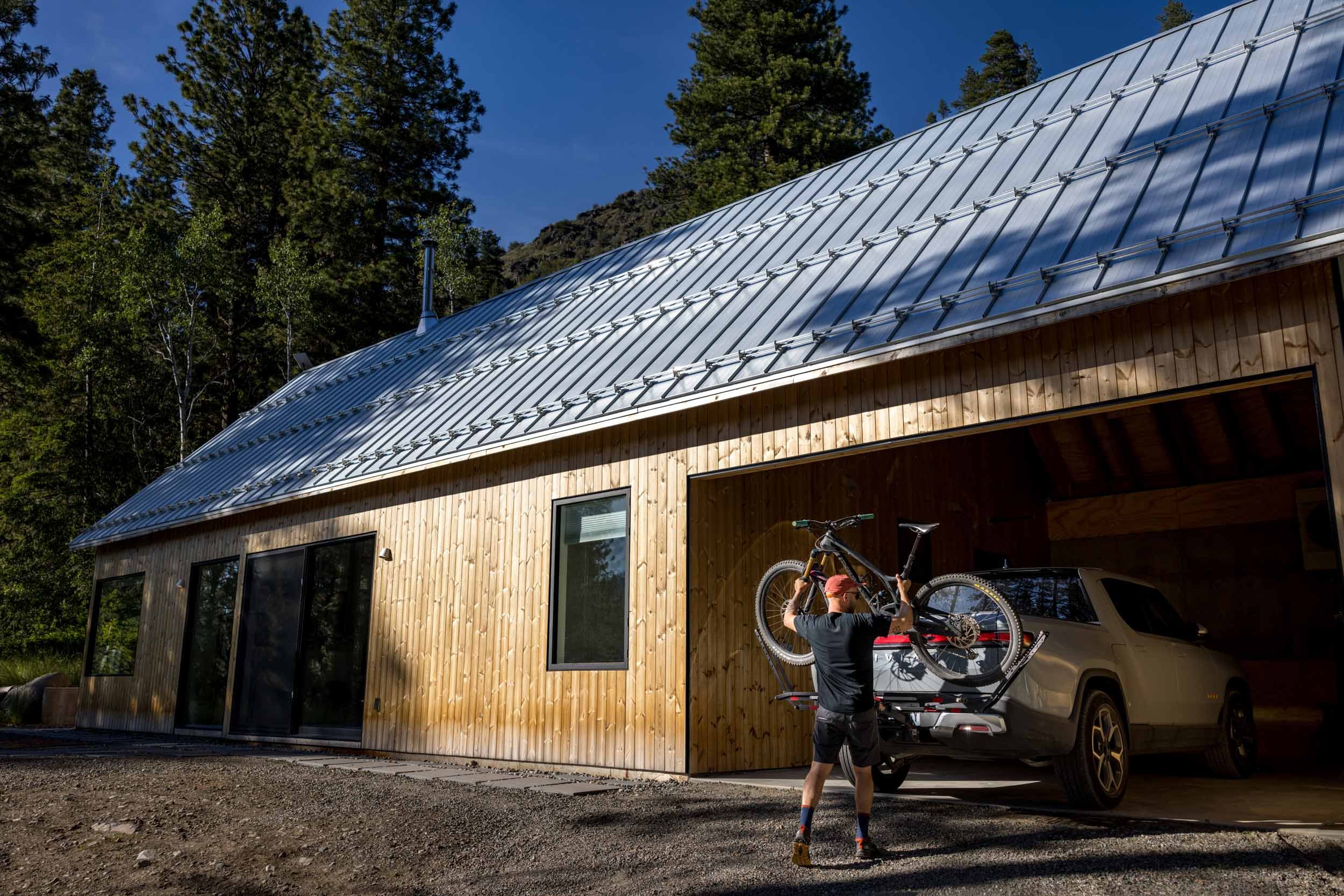 A person lifts a bicycle onto a rack attached to a parked car in front of a modern wooden house with a metal roof, surrounded by trees under a clear blue sky.