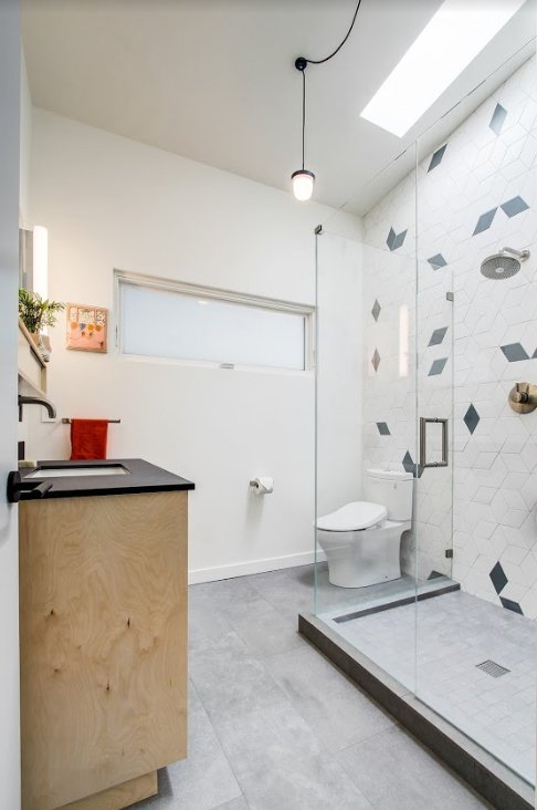 Modern bathroom with a glass shower, white geometric tile design, skylight, toilet, wooden vanity with black countertop, and minimalist decor.