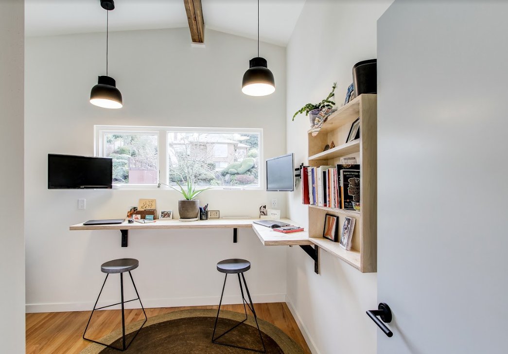 Modern home office with wall-mounted desk, two stools, hanging lights, bookshelves, and windowsill plant.