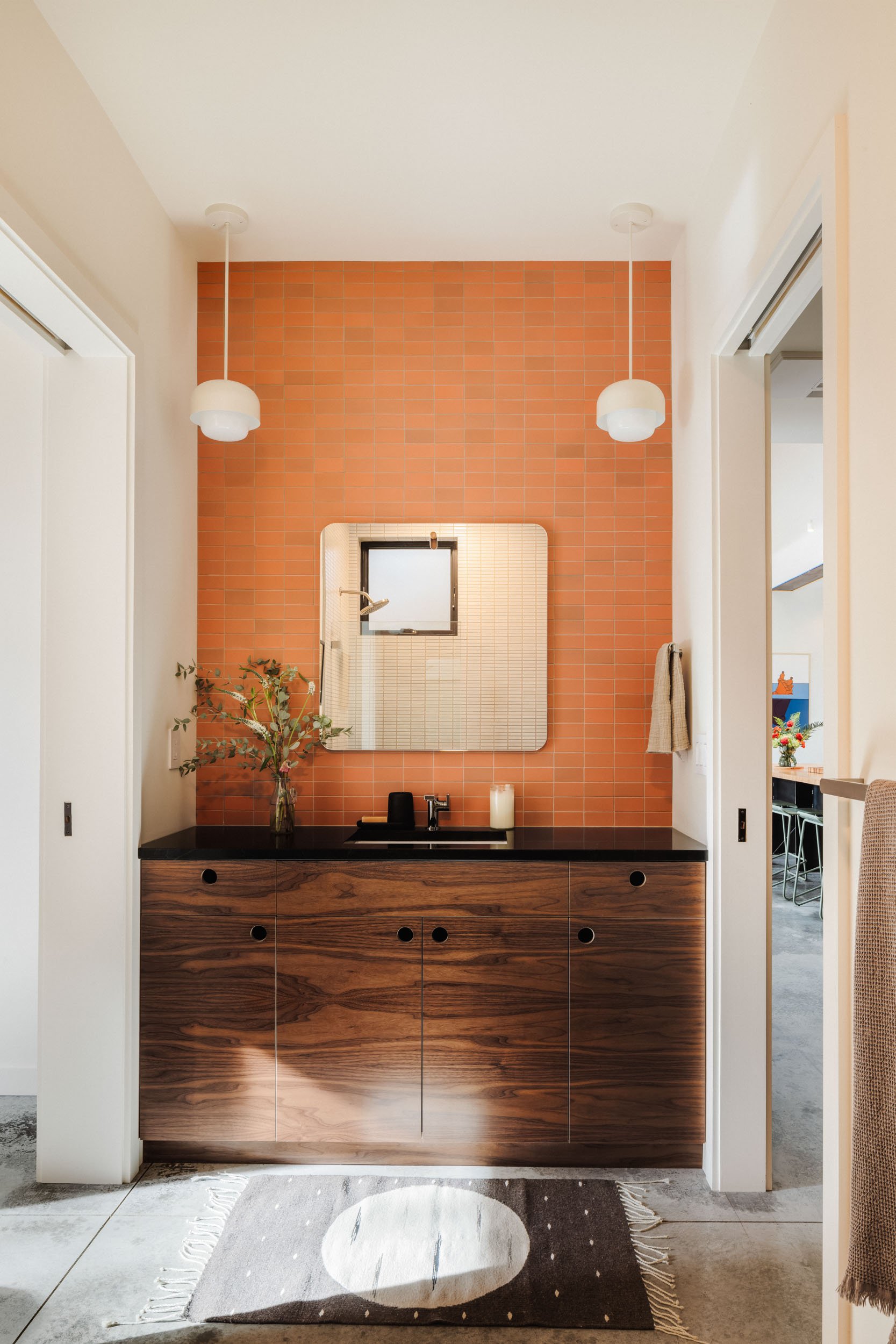 A modern bathroom interior featuring a wooden vanity with black countertop, a small rectangular mirror, and orange tiled backsplash. The countertop has a sink, a black soap dispenser, and a candle. Two hanging pendant lights flank the mirror. A plant