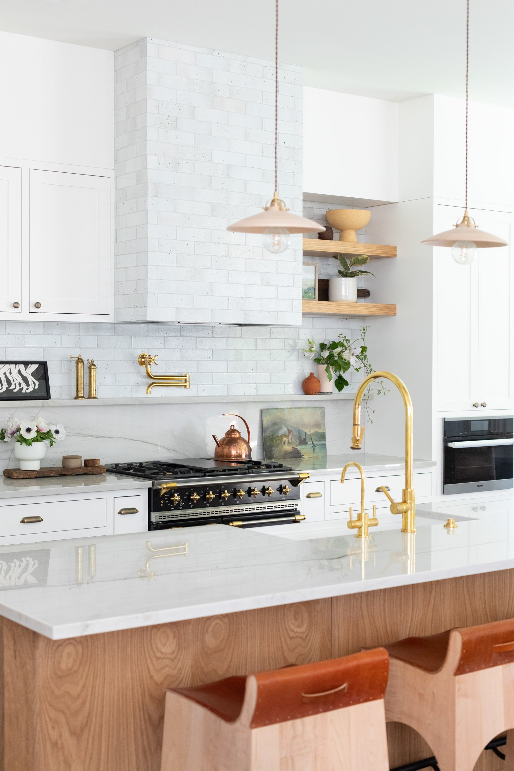 Modern kitchen with white cabinets, a marble countertop, and gold fixtures. Features a large black and gold stove, white subway tile backsplash, and open wooden shelves with plants and decor. Two pendant lights hang over the island with wooden stools