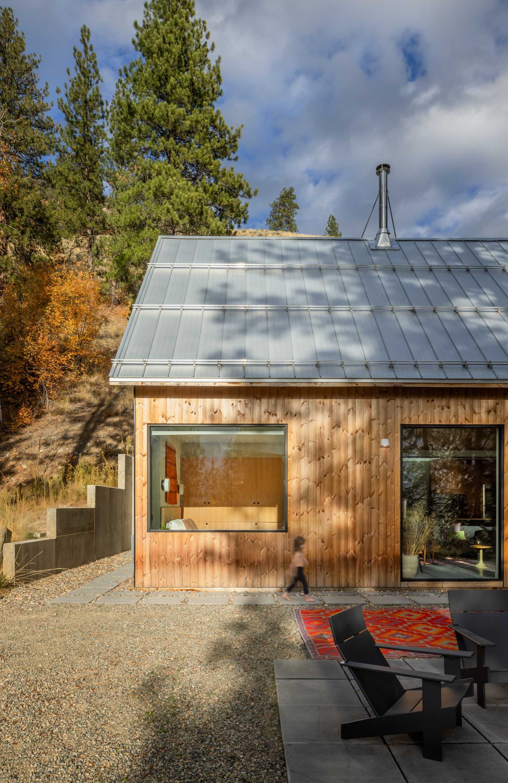 Modern wooden house with sloped metal roof and large windows; outdoor seating area with black chairs; surrounded by trees and autumn foliage.