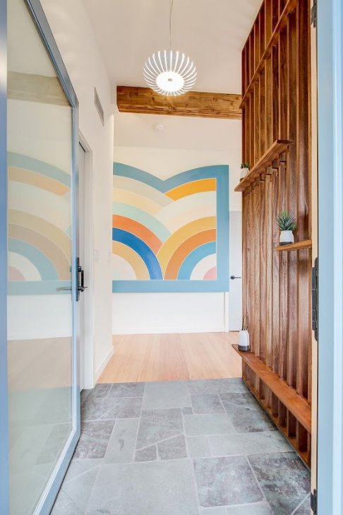 Entryway with stone floor and colorful geometric wall art, featuring wooden slatted partition and potted plants, illuminated by a modern pendant light.