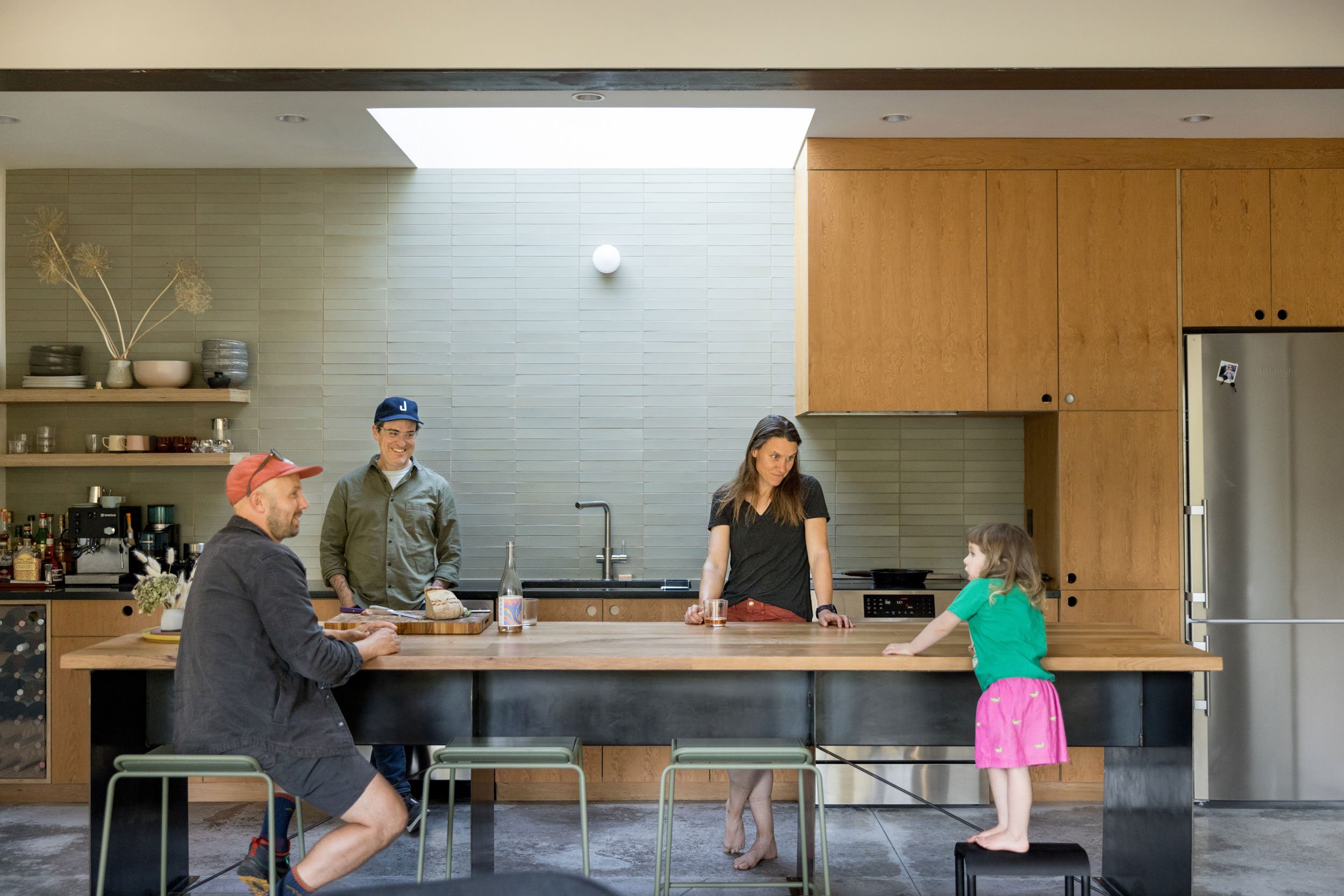 Family gathering in a modern kitchen with wooden cabinets and a large island. A man in a red cap sits at the island, another man stands near the sink, a woman and a young girl stand on the other side. Shelves hold plates and glassware.