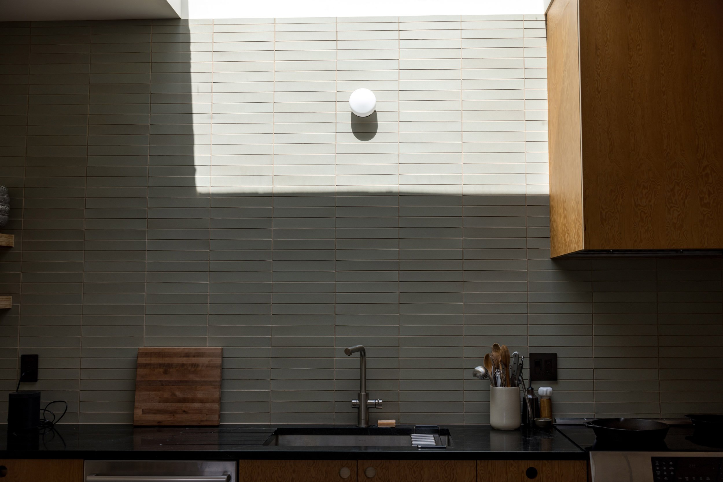 Modern kitchen with dark countertop, stainless steel faucet and black sink, tile backsplash, wooden cutting board, cutlery holder, and wooden cabinet.