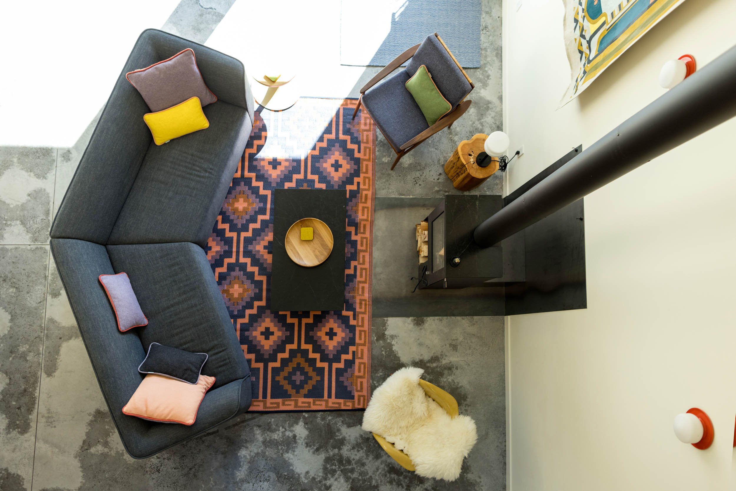 Overhead view of a modern living room with a gray L-shaped sofa, colorful cushions, geometric patterned rug, armchair with cushion, coffee table with tray, a wood-burning stove, and a fluffy chair.