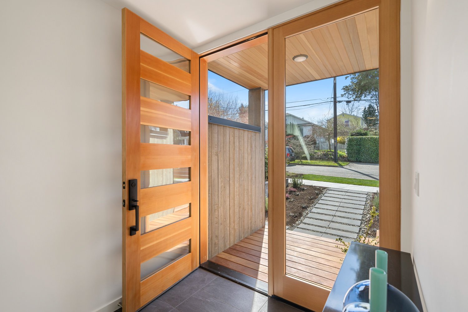 Open modern wooden front door with glass panels, leading to a porch and exterior pathway, with a view of residential street and greenery.