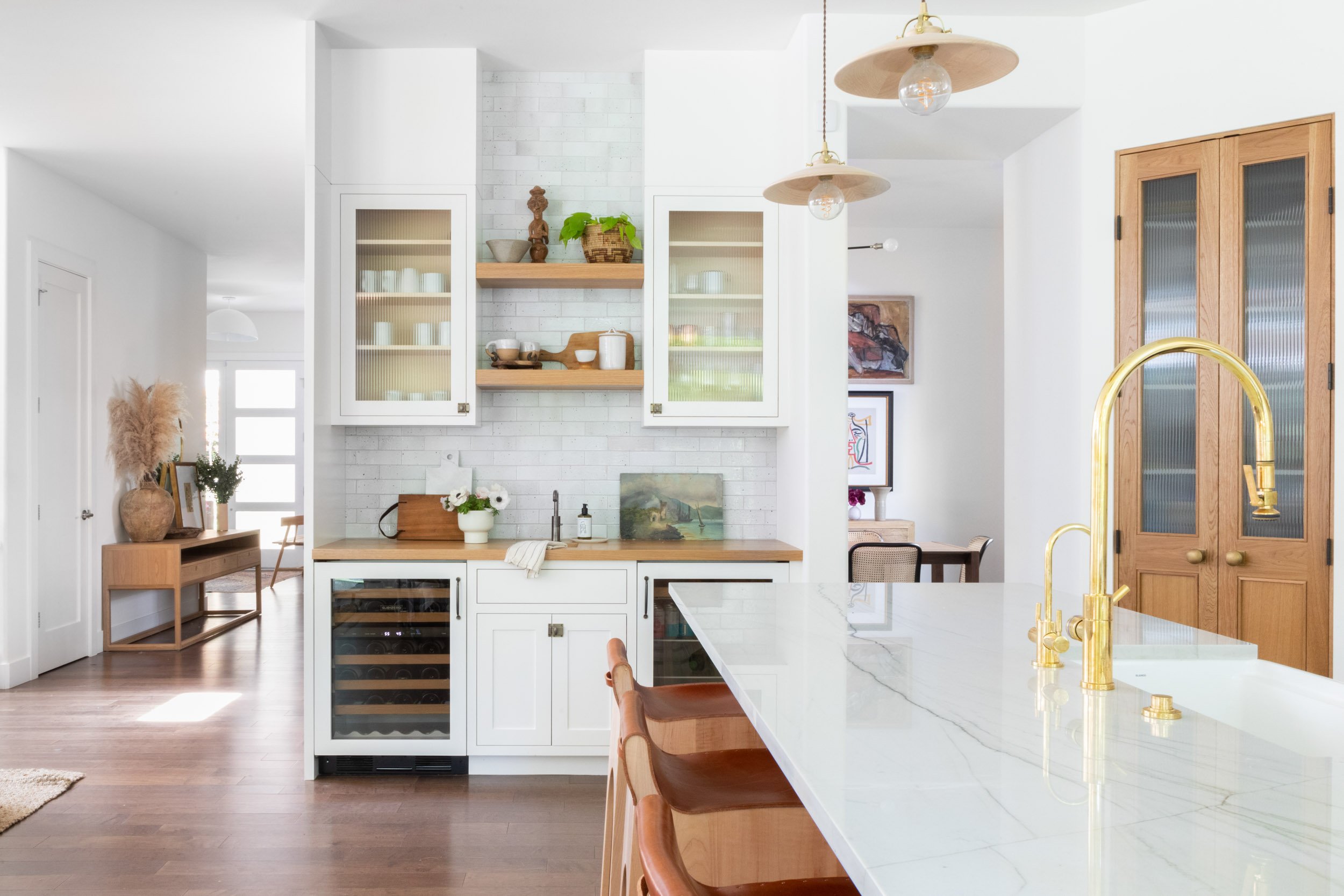 Modern kitchen with white cabinets, glass doors, wooden shelves, wine cooler, marble countertop, gold faucet, pendant lights, and wooden accents.