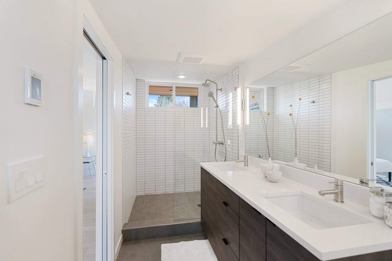 Modern bathroom with double vanity, large mirror, and walk-in shower featuring white tiles and minimalistic decor.