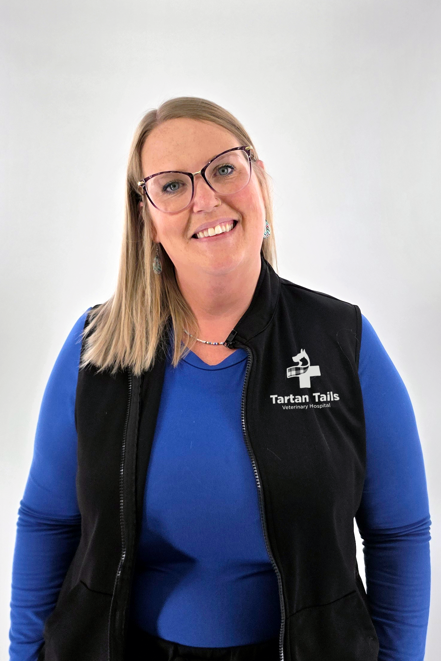 A smiling woman wearing glasses, blue scrubs with a veterinary hospital logo, and a stethoscope around her neck.