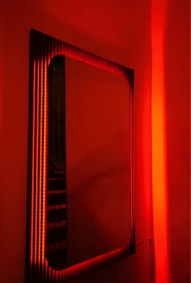 A large rectangular mirror with a frame illuminated by red LED lights, mounted on a red-painted wall.