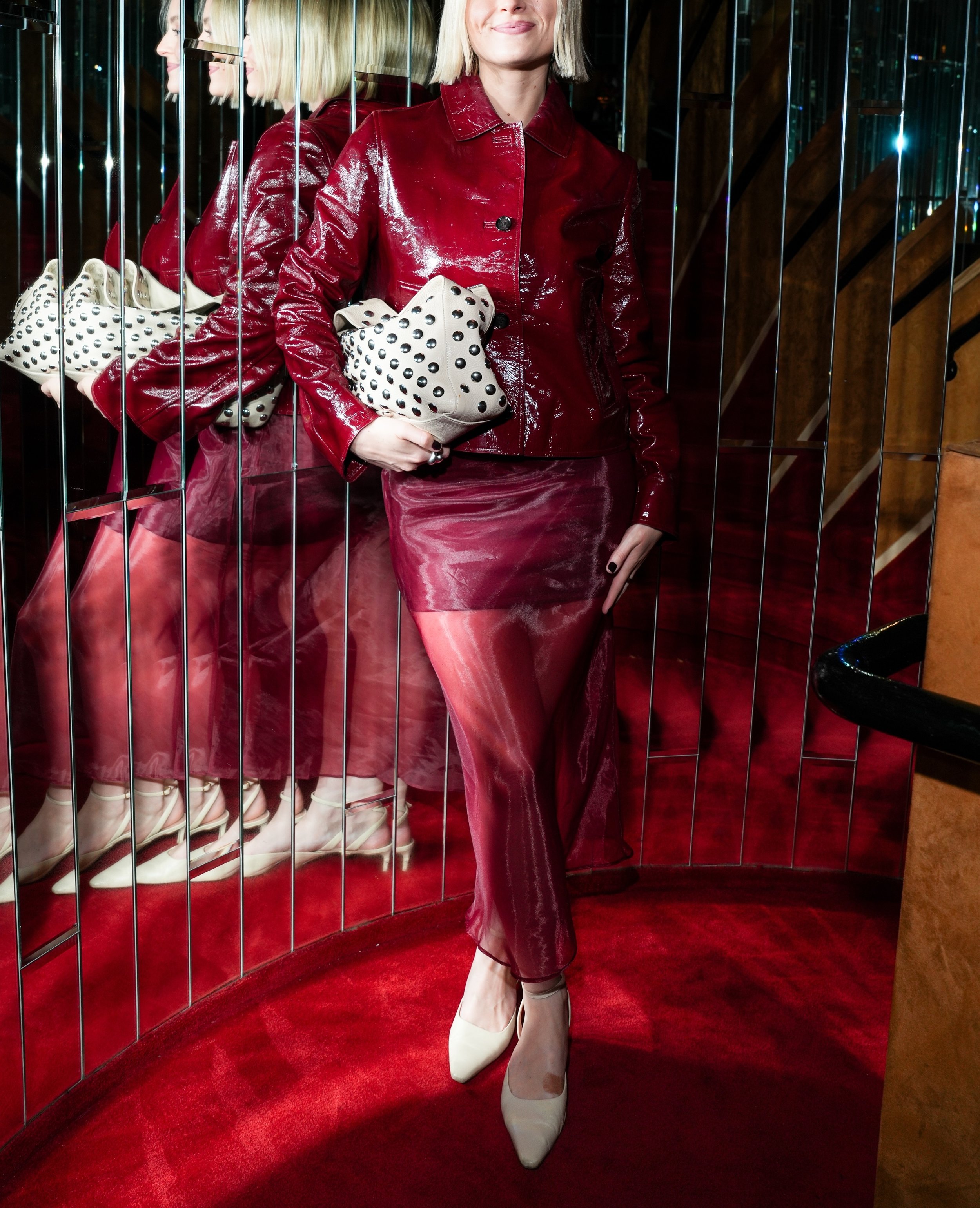 Woman in a shiny red jacket and skirt with a white clutch posing in mirrored walls at a chic Soho NYC cocktail bar