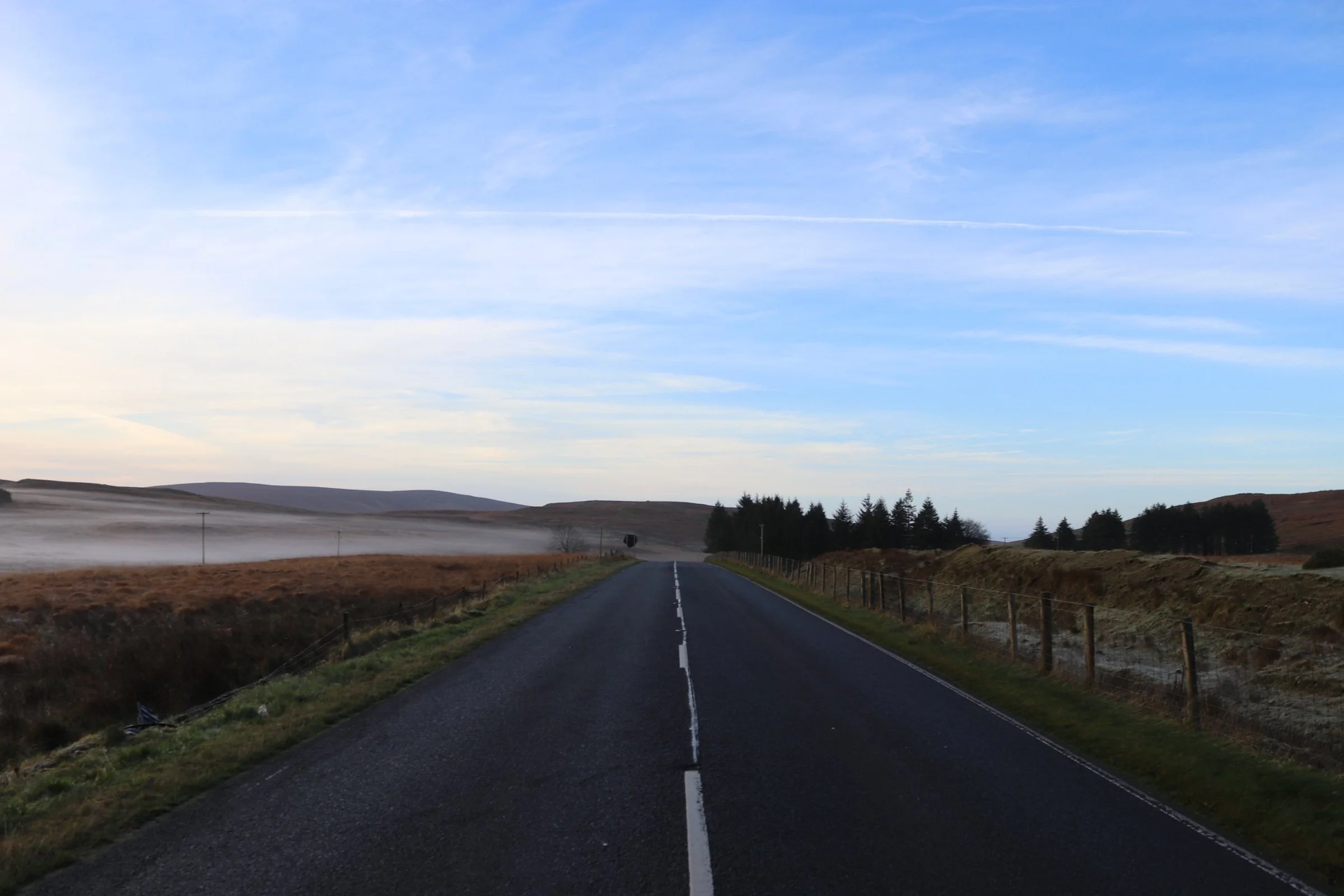 A straight rural road stretching into the distance with grassy fields and sparse trees on either side, under a partly cloudy sky.