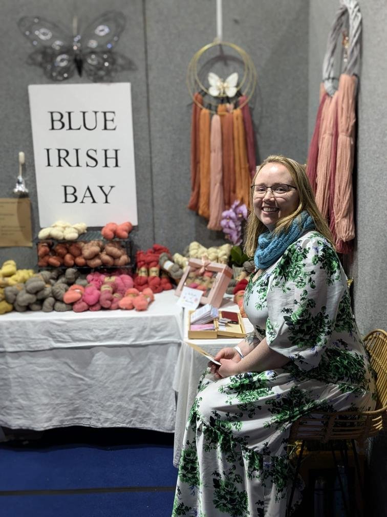 Irish woman with glasses and blonde hair, smiling and sitting on a wicker chair at a craft stall. The stall has a large sign reading 'Blue Irish Bay,' colorful yarn skeins she has hand dyed for sale, and a display of yarn hanging from the wall.