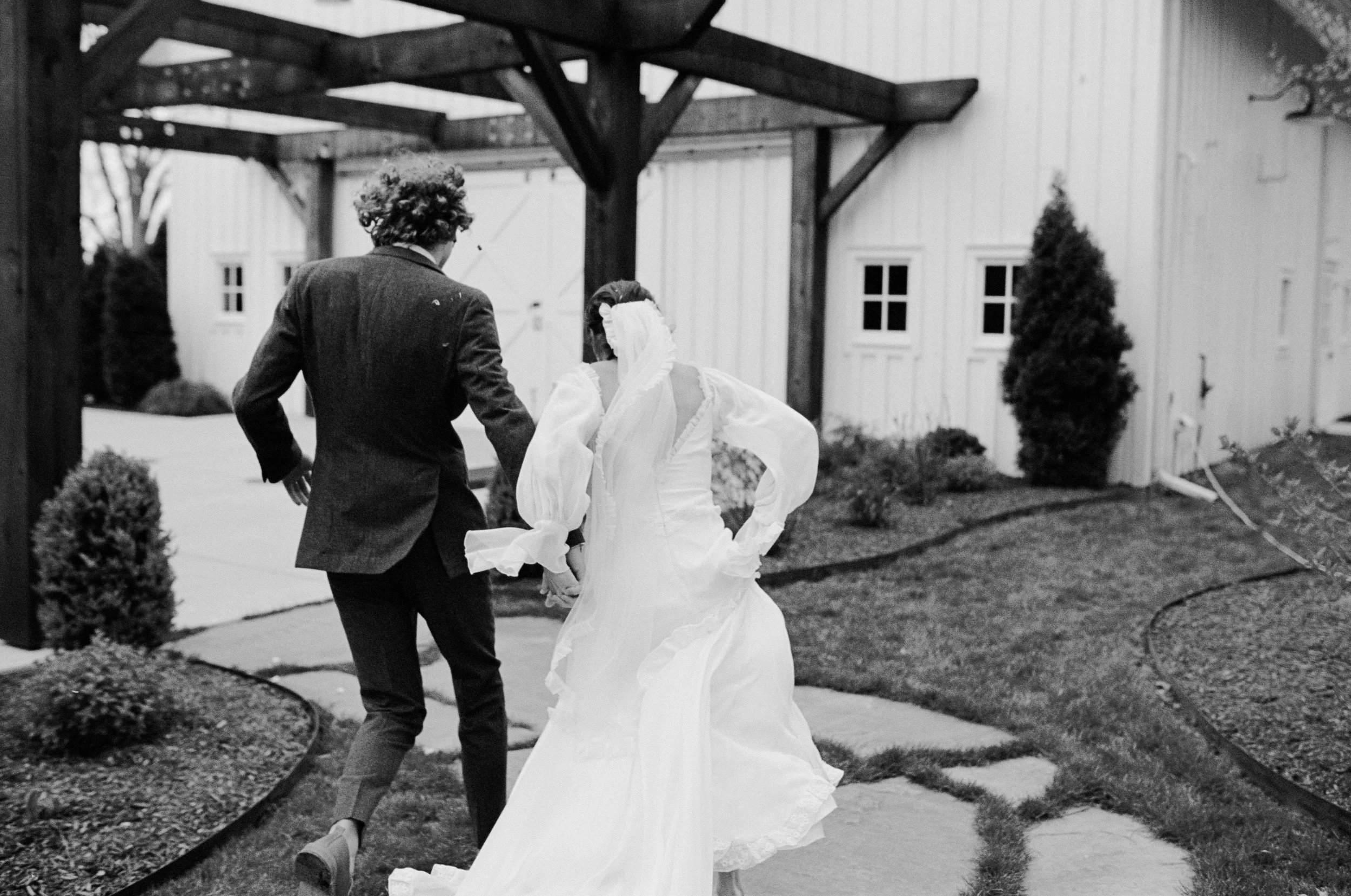 A black and white photo of a bride in a wedding dress and a man in a suit holding hands, walking outdoors near a white building with trees and shrubs in the background.
