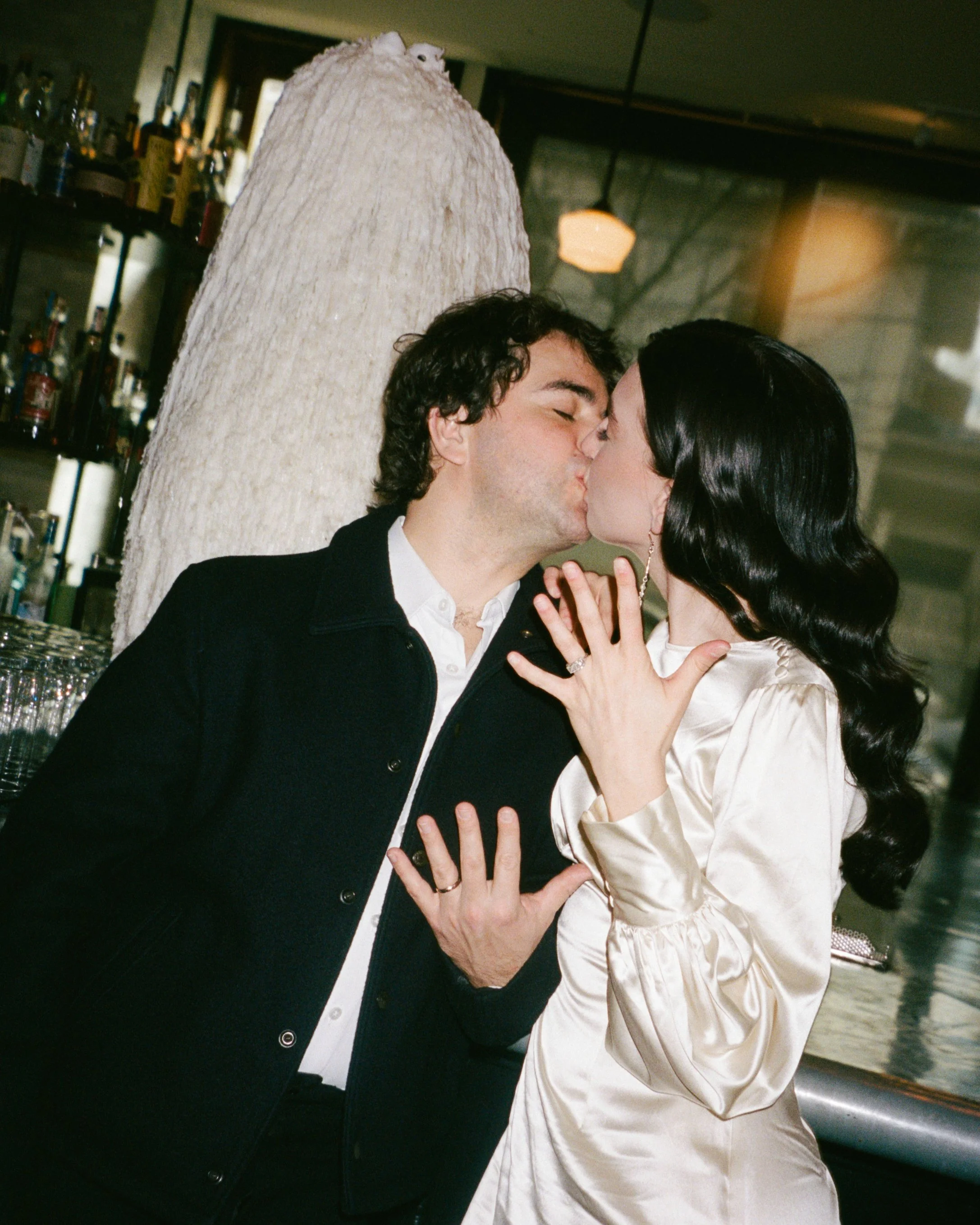 A man and woman kissing in a bar, with the woman showing her hand with an engagement ring.