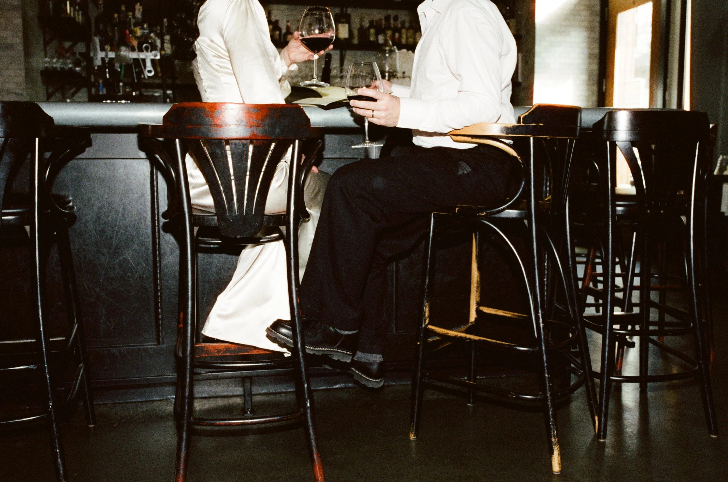 Two people sitting at a bar with glasses of red wine, having a conversation in a dimly lit pub or restaurant.