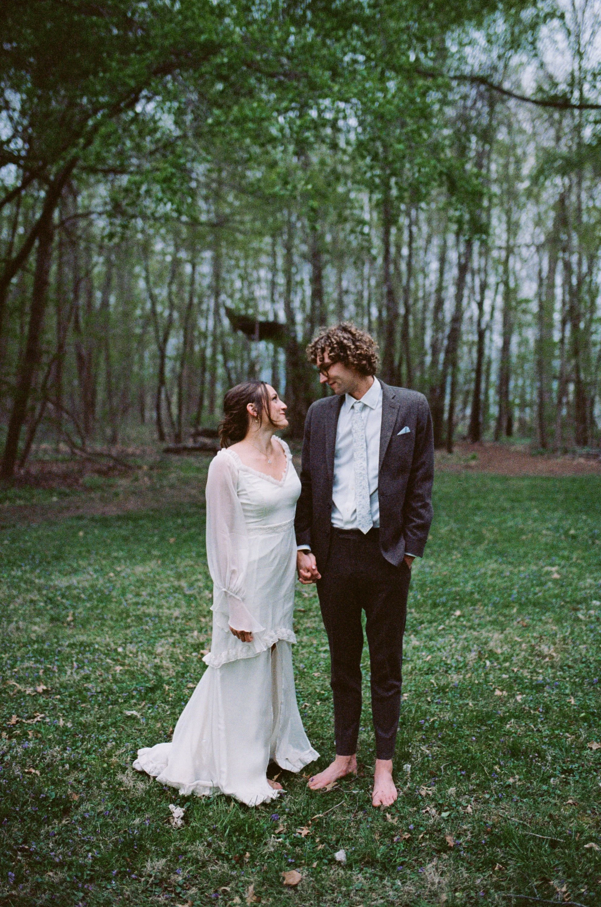 A couple stands barefoot on a grassy forest clearing, holding hands while gazing into each other's eyes. The woman wears a white bohemian-style dress with long sleeves, and the man wears a dark suit with a white shirt, no tie, and a light blue pocket