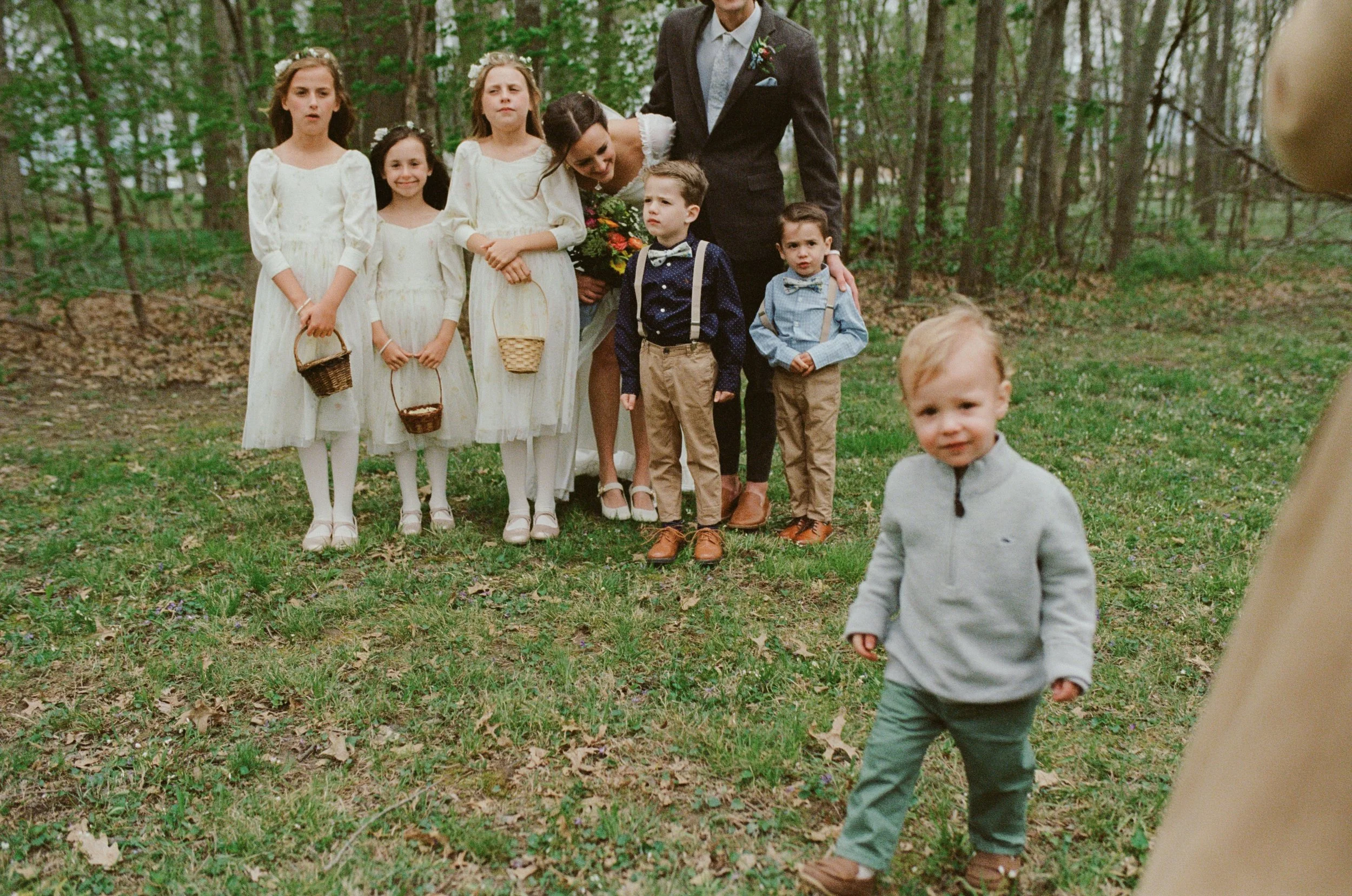 A group of children dressed in vintage-style clothing standing outdoors in a wooded area, with a woman and a man dressed in formal attire among them. One boy is running toward the camera in the foreground.