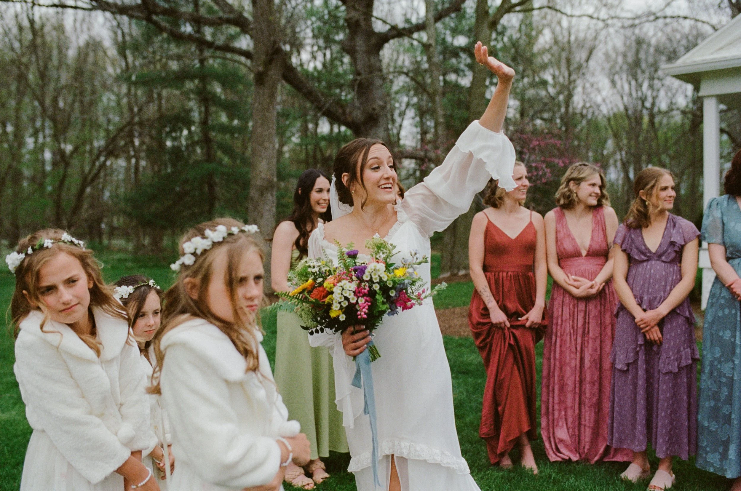 A bride holding a bouquet of colorful flowers smiling and raising her arm during an outdoor wedding ceremony with bridesmaids and flower girls in colorful dresses and flower crowns, surrounded by trees.