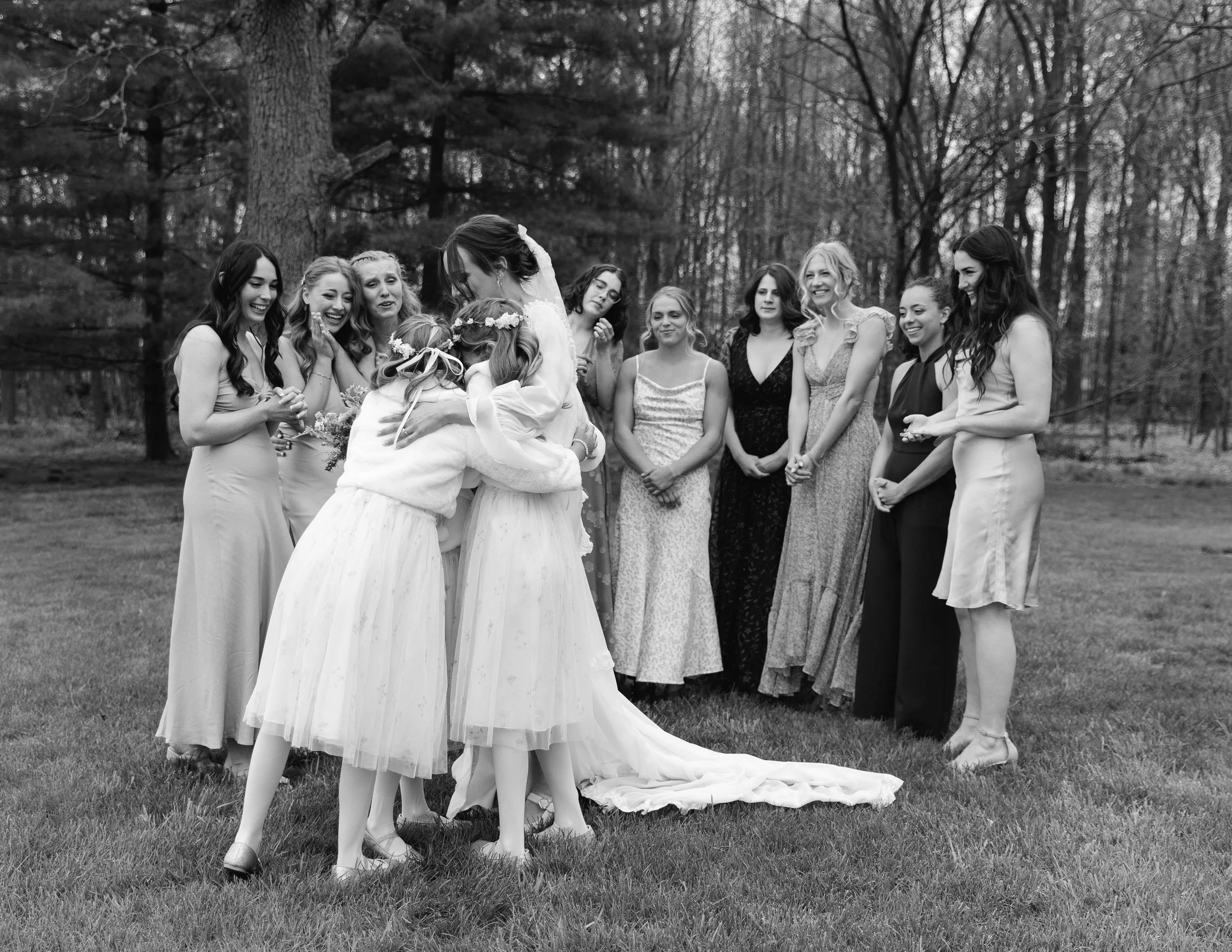 A group of women gathered outdoors in a wooded area, some dressed in formal wear, hugging and smiling during a wedding celebration.