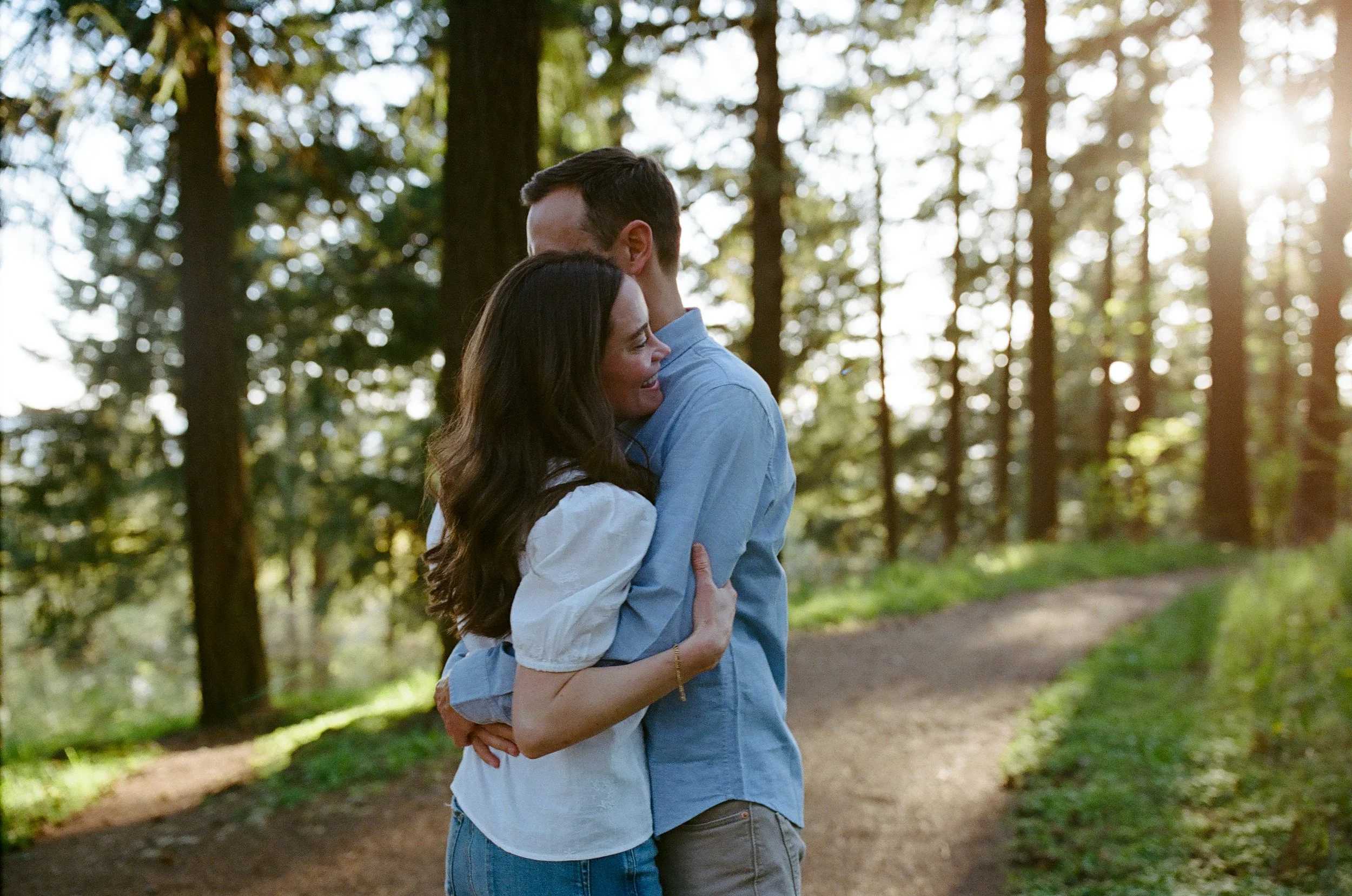 A couple hugging in a forested area with sunlight filtering through the trees.
