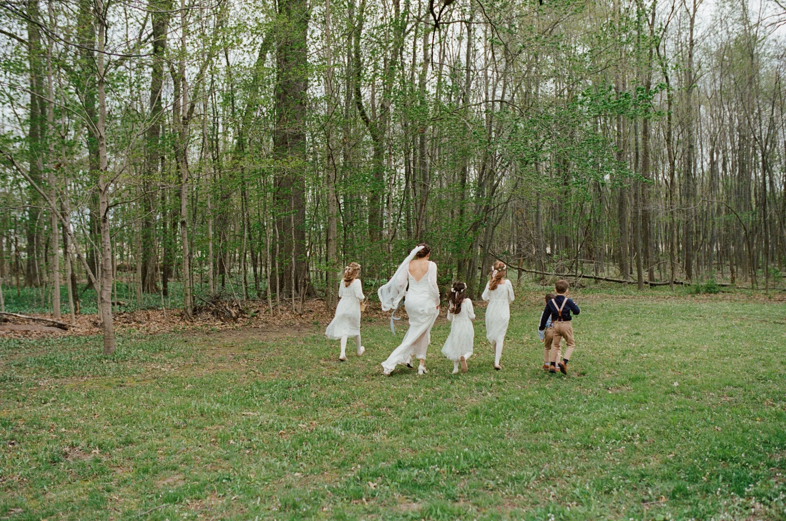 A bride and five children walking in a forest clearing during daytime. The bride wears a white dress and veil, and the children are dressed in white and tan, with one boy in suspenders. The background consists of trees and greenery.