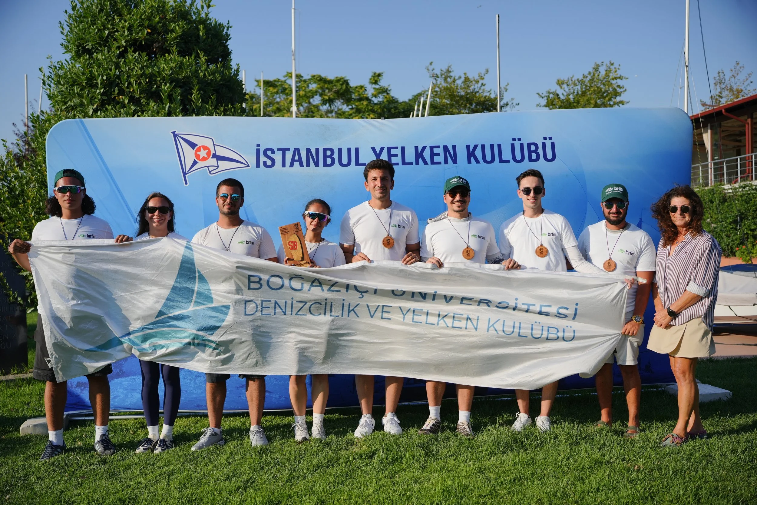 İstanbul Yelken Kulübü üyeleri, açık havada, güneşli bir gün, gökyüzü mavi, arka planda yeşil ağaçlar ve bazı yapılar ile birlikte, üzerinde İstanbul Yelken Kulübü yazılı büyük bir mavi pankart tutuyorlar, yanlarında bir bayan, hepsi sportif kıyafetler giymiş, arkadaş canlısı ve mutlu görünüyorlar.