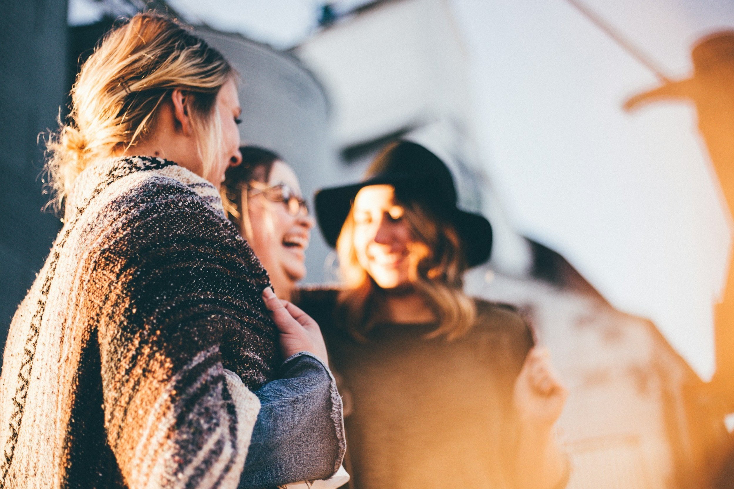 Tres mujeres riendo y sonriendo al estar juntas, una lleva sombrero negro y otra con gafas de sol, en un ambiente soleado.