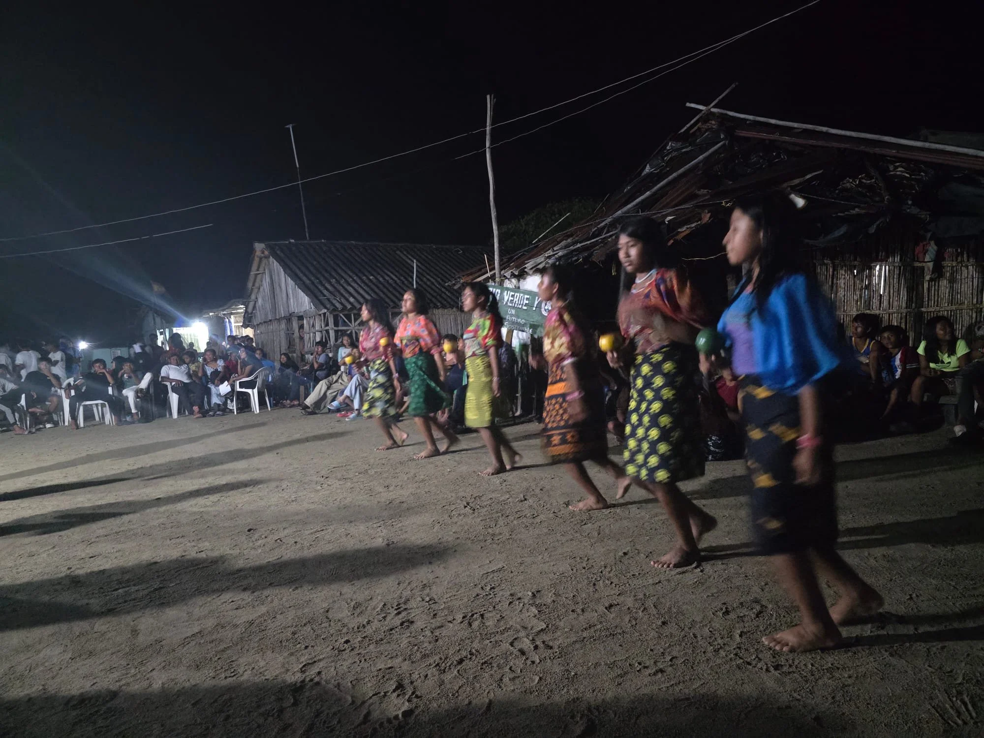 Group of women in colorful traditional dresses walking barefoot on dirt ground during night time event outside wooden huts, some sitting and some standing.