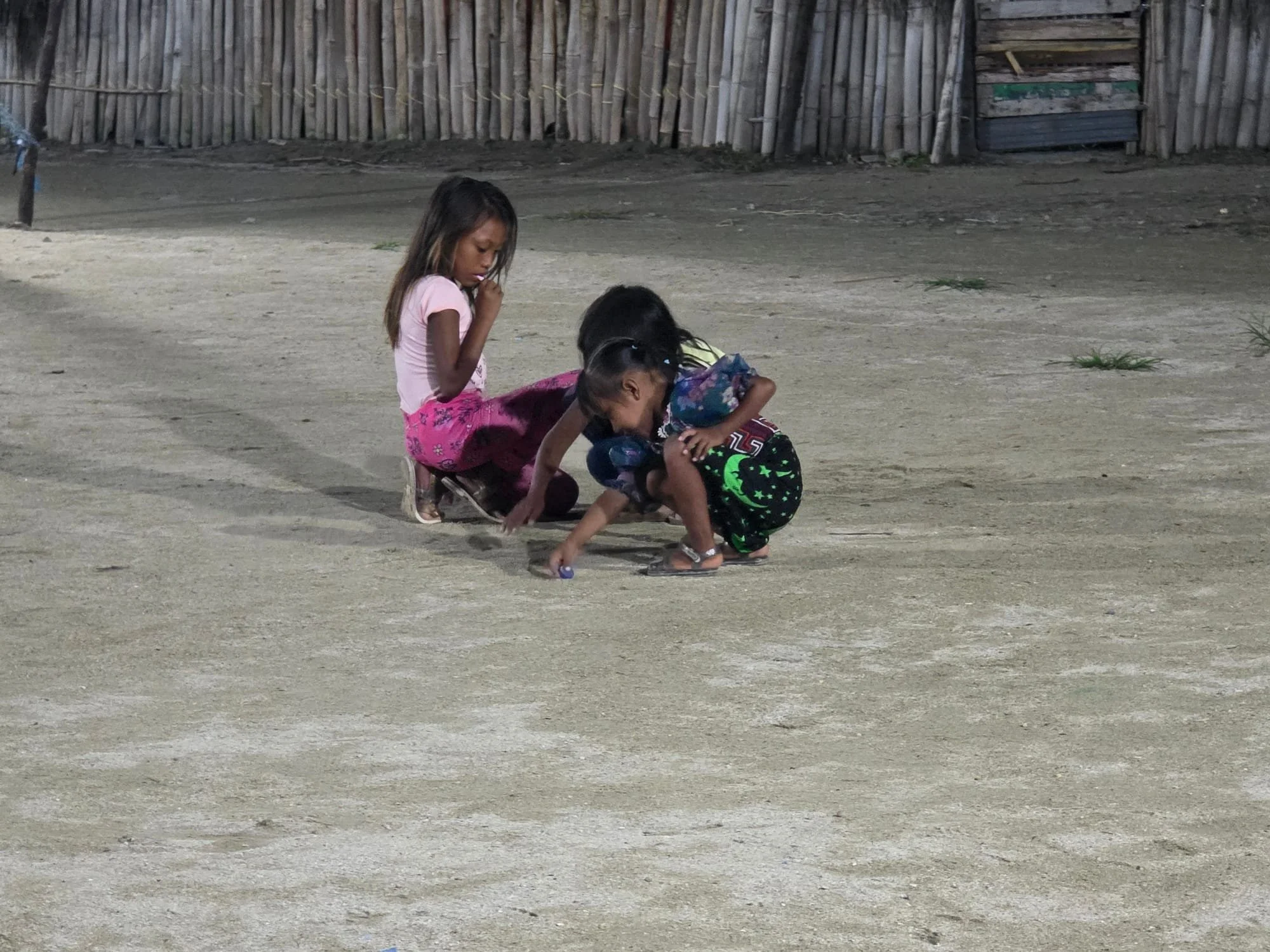 Three children playing on a dirt ground in front of a wooden fence, with one girl wearing a pink dress and two other children crouching and reaching towards the ground.