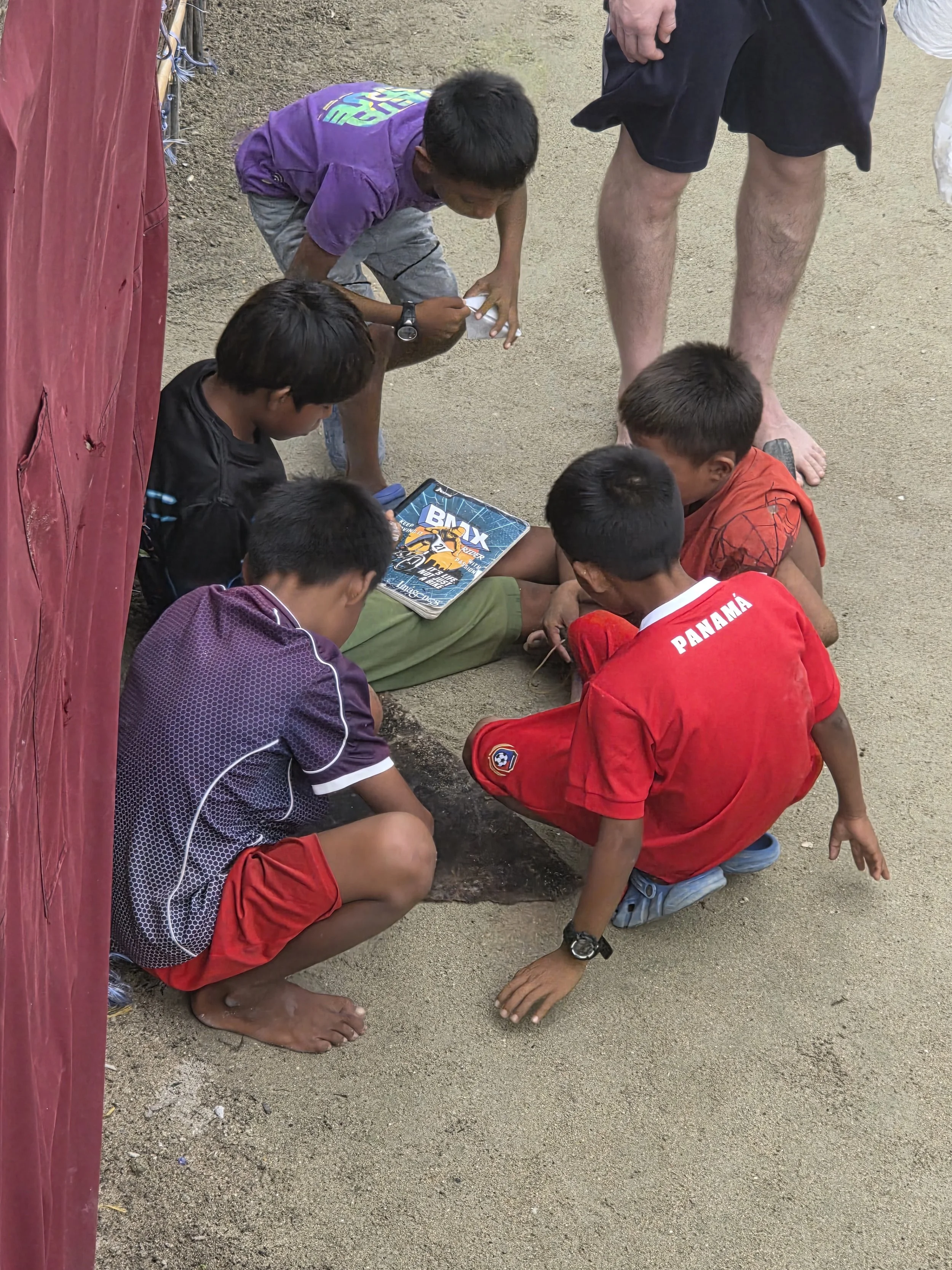 A group of children and one adult sitting and kneeling on the ground, focusing on a small hole in the dirt, with a tablet nearby showing a racing game.