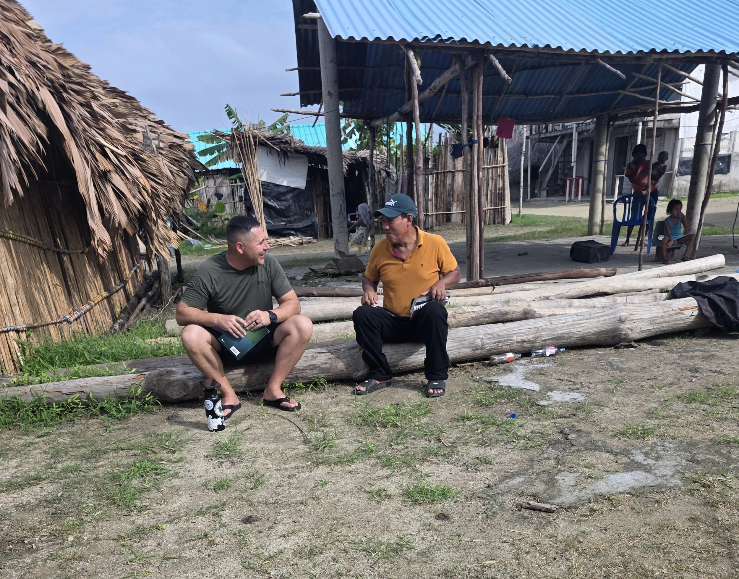 Two men sit outdoors talking in front of bamboo and thatch structures, with children and a woman nearby. It appears to be a rural village setting.