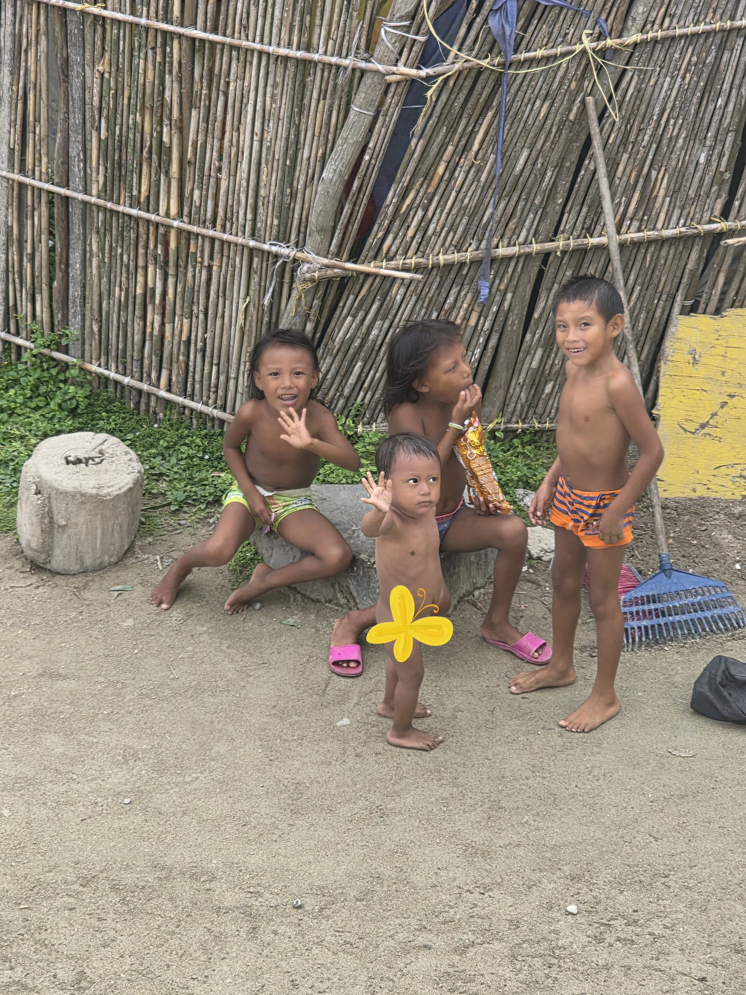 Four young children, three girls and one boy, are standing and sitting on a dirt ground in front of a bamboo and wooden fence. They are shirtless, with some wearing shorts and others wearing slippers. One girl is holding a snack while another girl an
