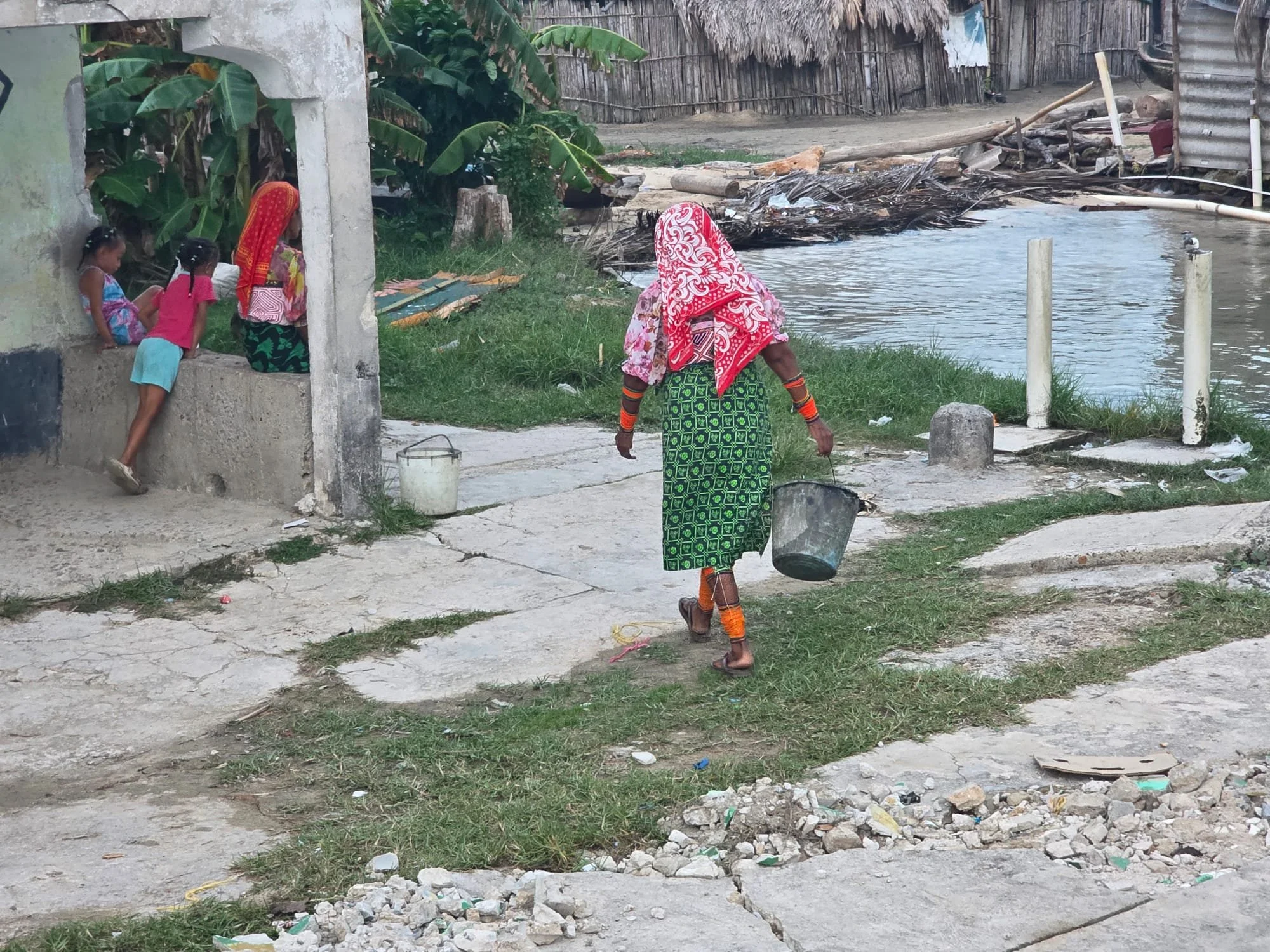 An elderly woman wearing a colorful patterned headscarf and skirt, carrying buckets, walking on a dirt path by a river. In the background, three children sit on a wall near green banana plants, with a worn wooden fence and a simple shelter nearby.