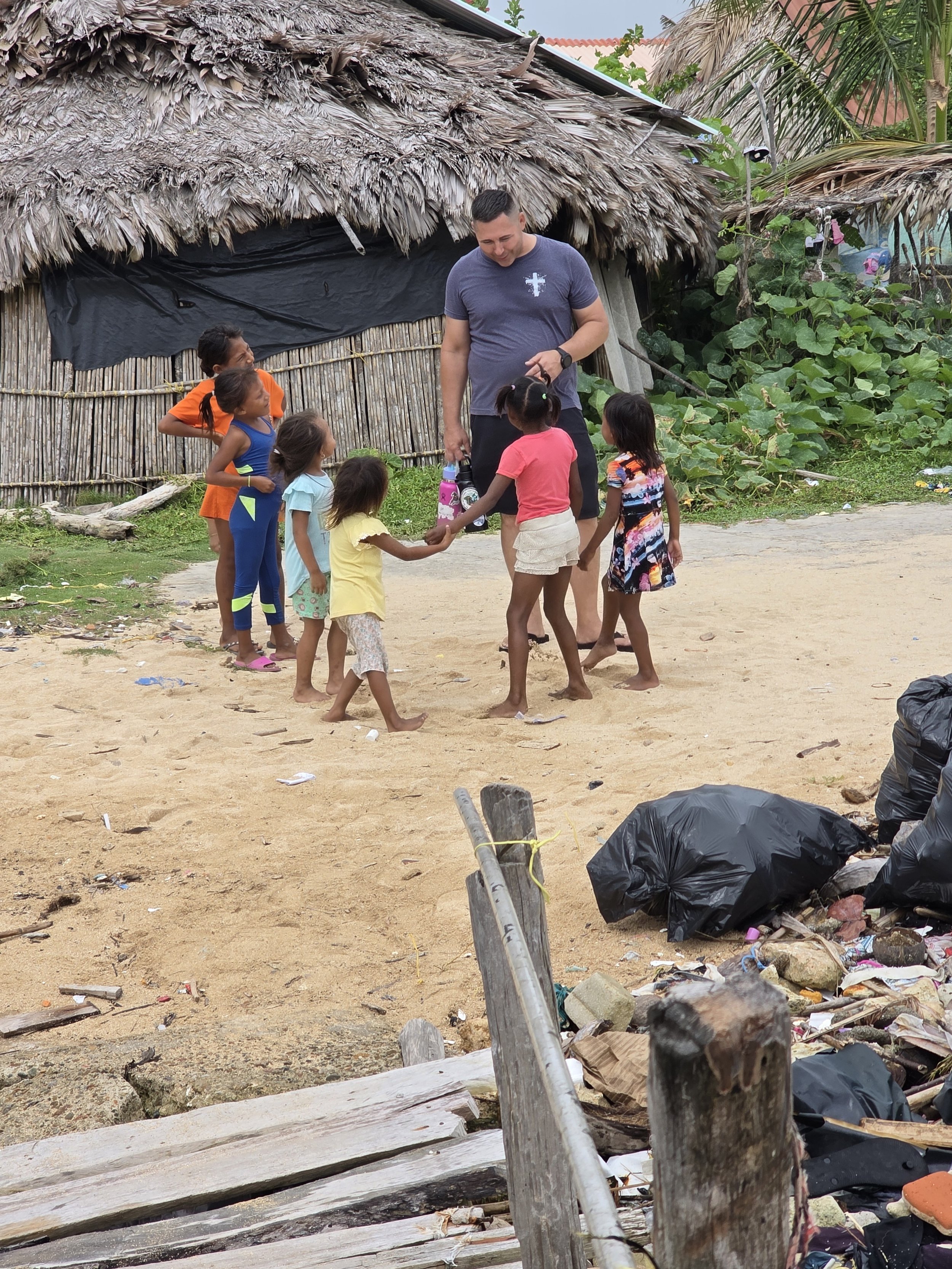 A man talking to five young girls in a rural outdoor setting, with a thatched roof building and lush greenery in the background.