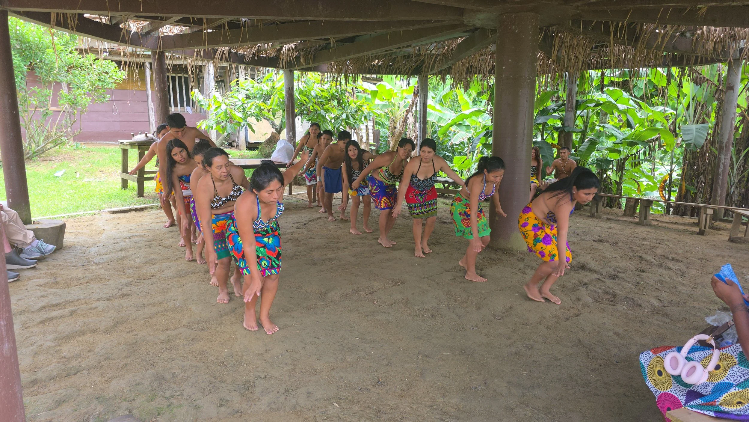 Group of people in traditional clothing participating in a cultural dance or activity under a thatched roof structure surrounded by lush greenery.