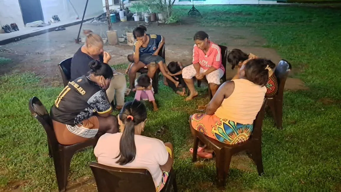 A group of women and children sitting in a circle outdoors during evening or nighttime, some with heads bowed and eyes closed, appearing to be praying or meditating.