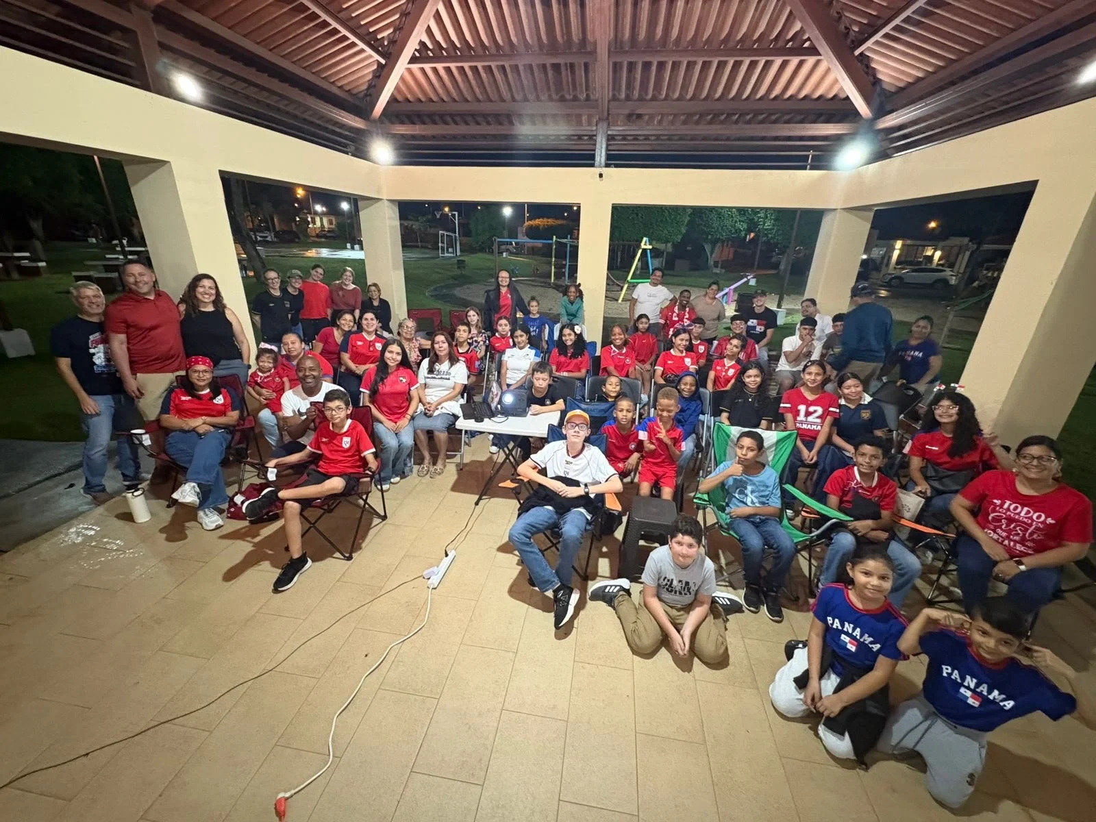 A large group of children and adults gathered together outdoors at night in a pavilion, some seated and some standing, for a group photo. Many are wearing sports jerseys, with a visible Panama and USA team shirts.