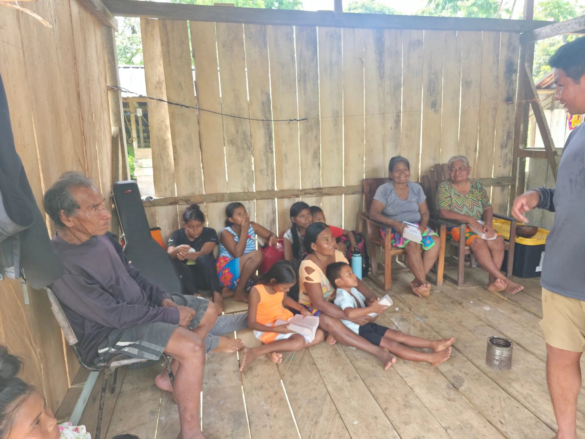 Group of children and elderly people sitting inside a wooden structure with unfinished walls, some holding notebooks, and a young man standing on the right side.