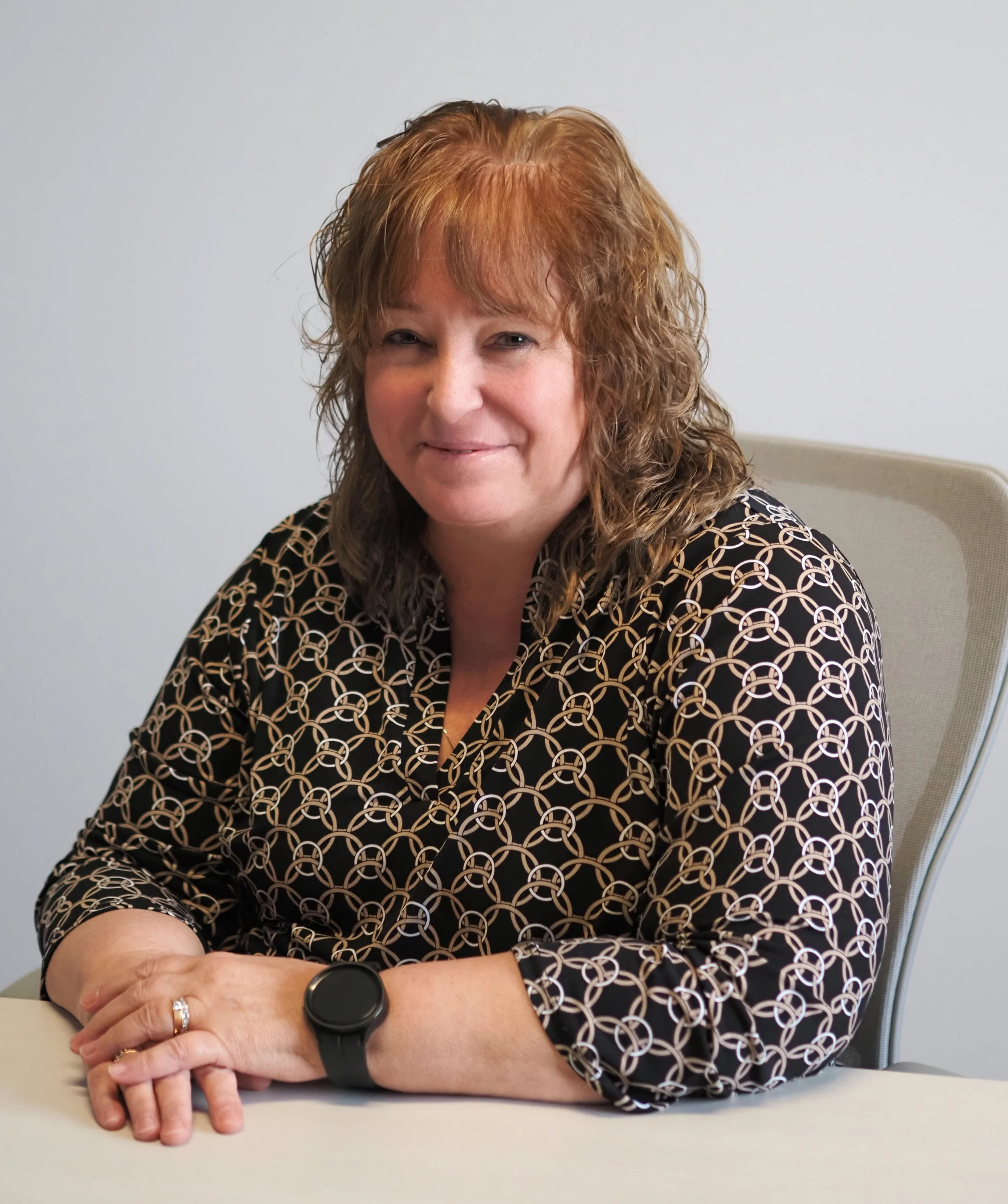 Sylvie Charron seated at a table, wearing a patterned blouse and a wristwatch, smiling.