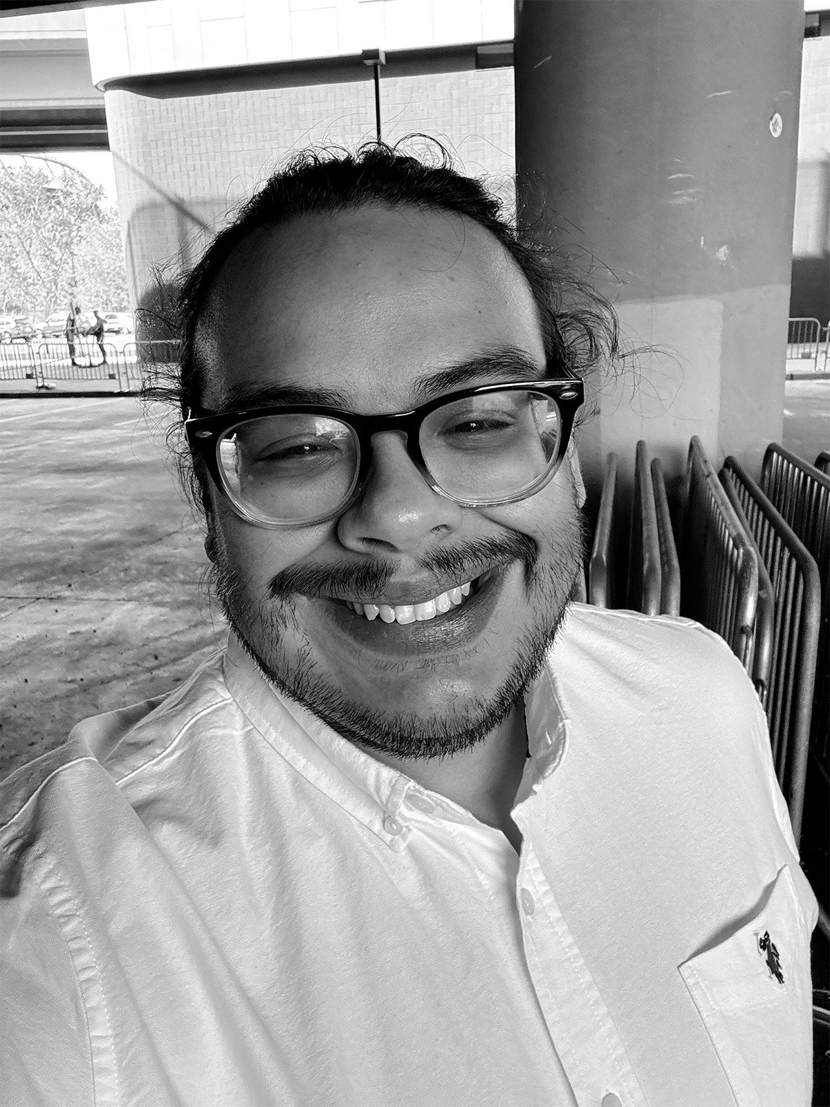A smiling person with glasses and facial hair, wearing a button-up shirt, outdoors with metal chairs and a pole behind them.