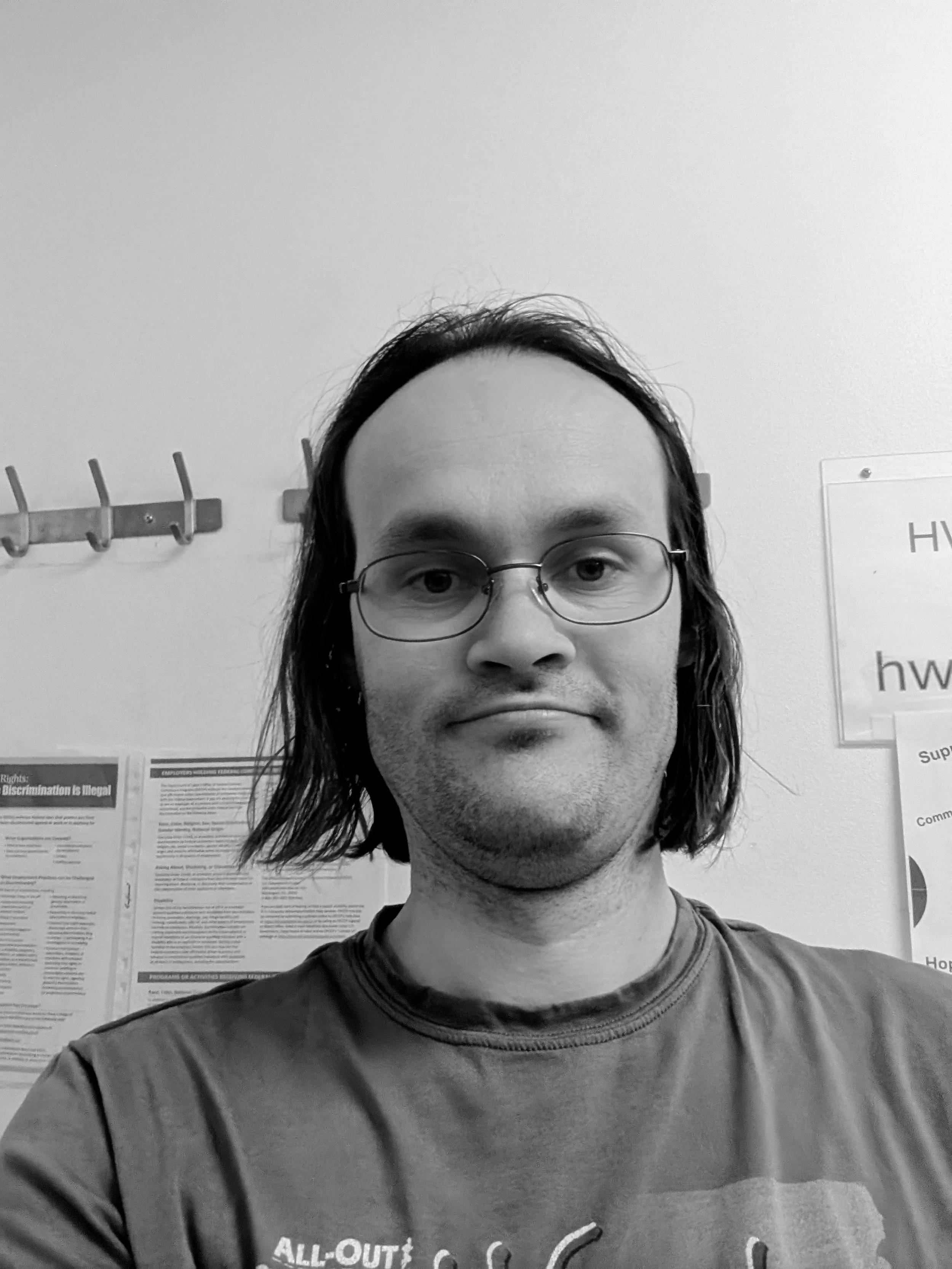 Black and white photo of a man with long hair and glasses, outdoors in front of a wall with informational posters and a coat rack, looking directly at the camera.