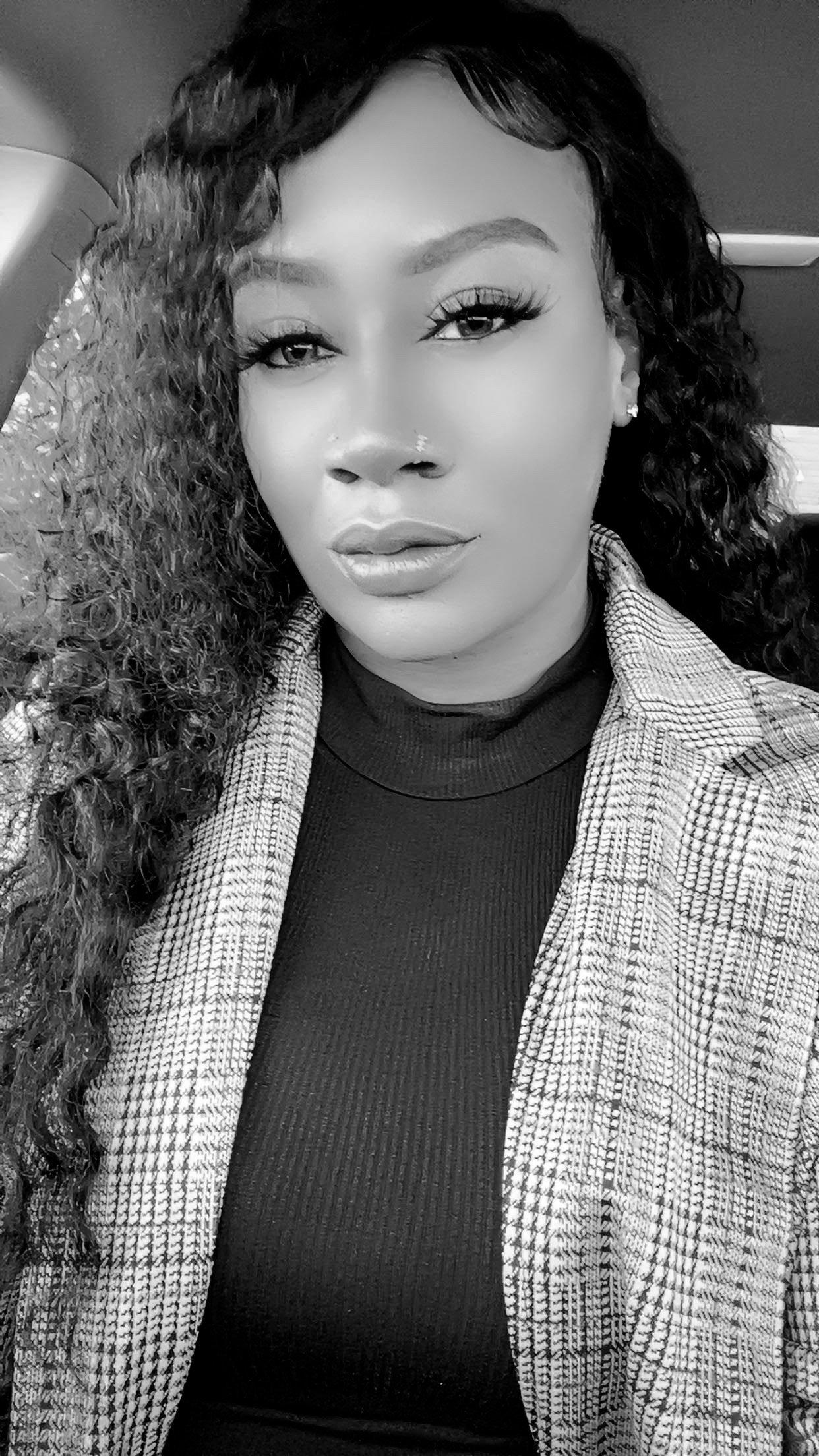 A woman with curly hair, wearing earrings and a checked blazer, looking at the camera in a close-up black and white portrait.