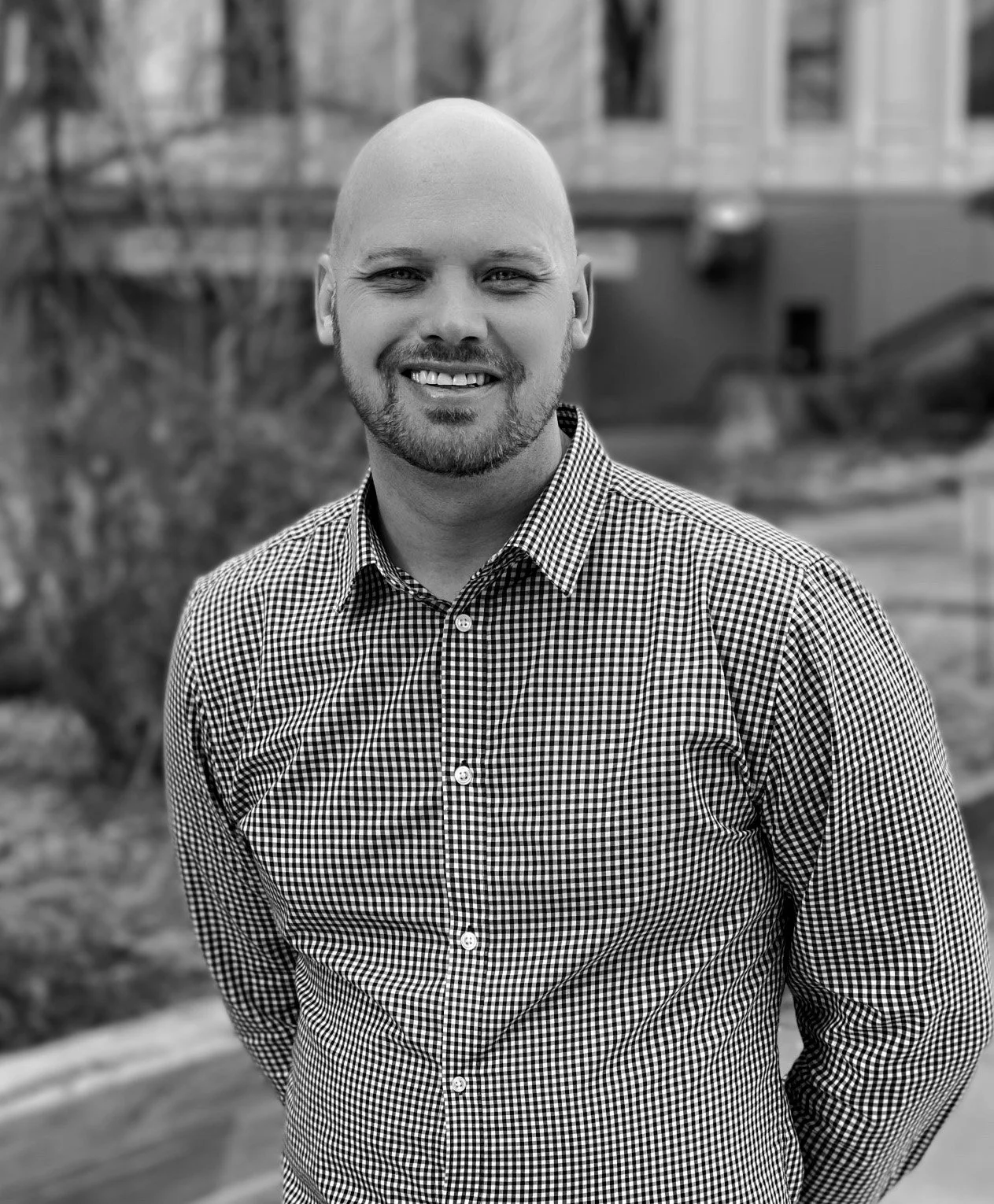 A black and white portrait of a smiling man with a beard, wearing a checkered shirt, outdoors with blurred buildings and trees in the background.