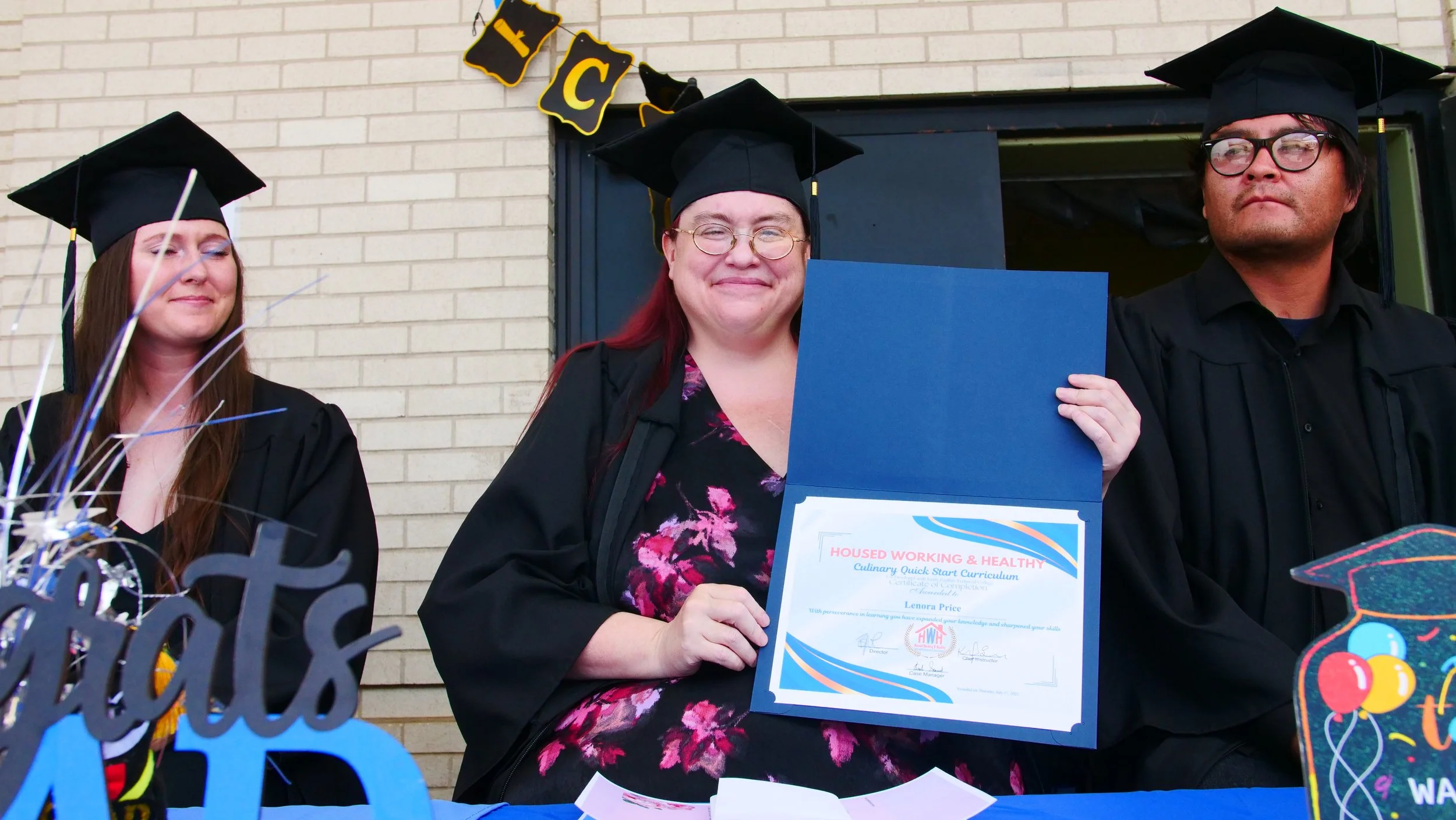 Three graduates in caps and gowns standing outdoors, with a woman in the center holding a certificate, smiling. Colorful decorations and a brick wall in the background.